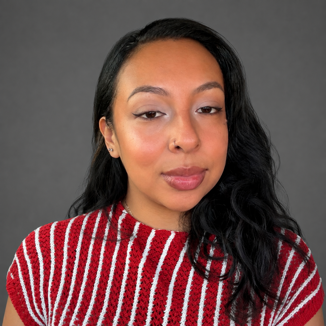 Close-up portrait of a young woman with braided hair, wearing gold hoop earrings and a beige shirt, looking at the camera against a plain gray background.