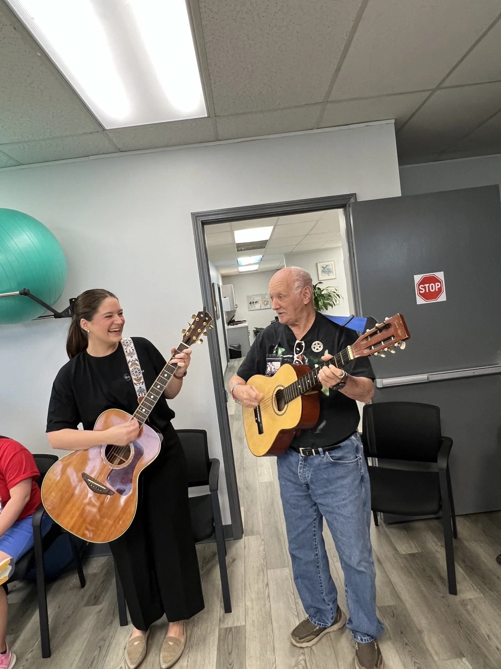 A young woman and an elderly man playing guitars and singing together indoors, smiling and enjoying the moment.