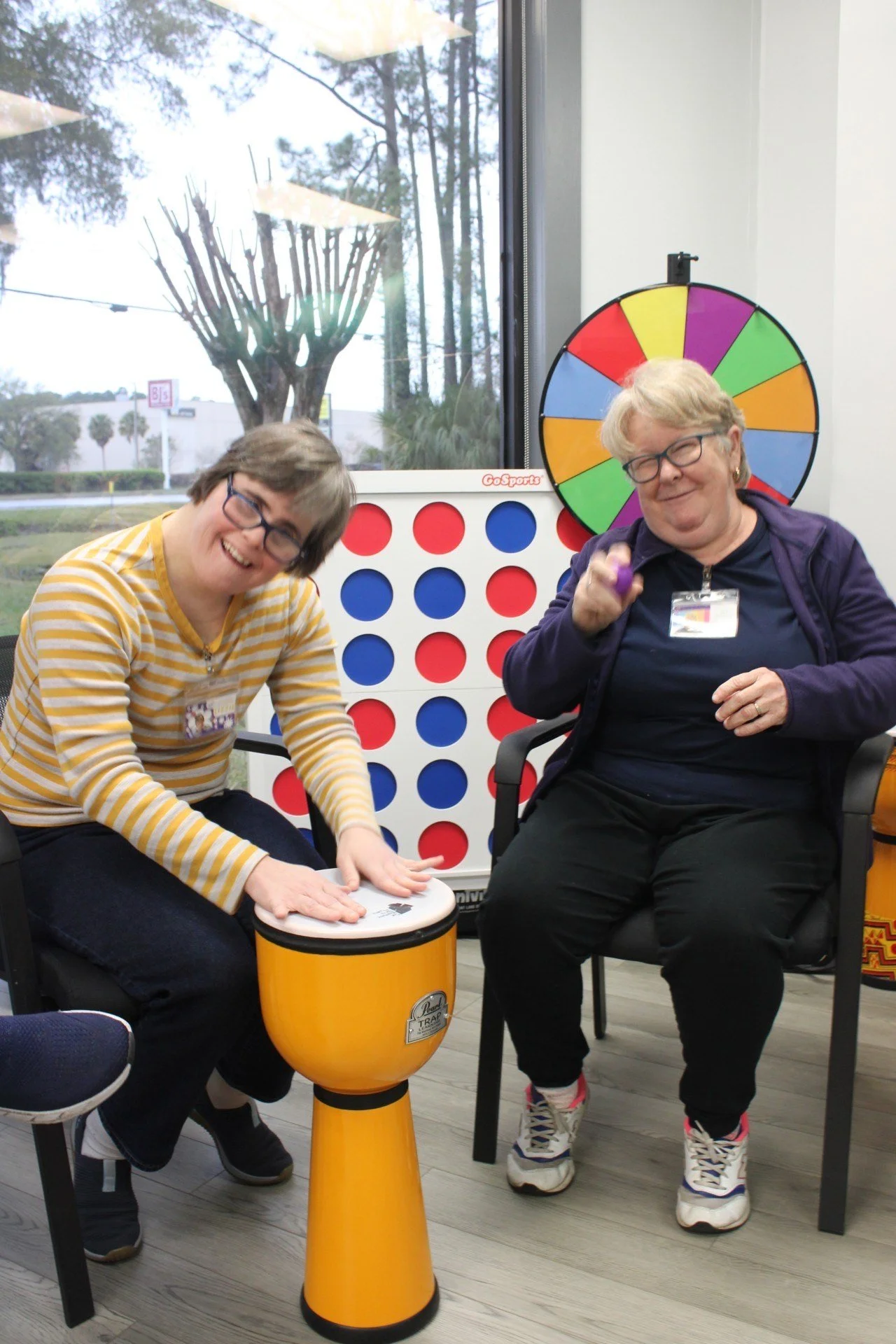 Two women sitting in chairs, playing with small percussion instruments next to a large Connect Four game and a colorful spinning wheel in a room with a window showing trees outside.