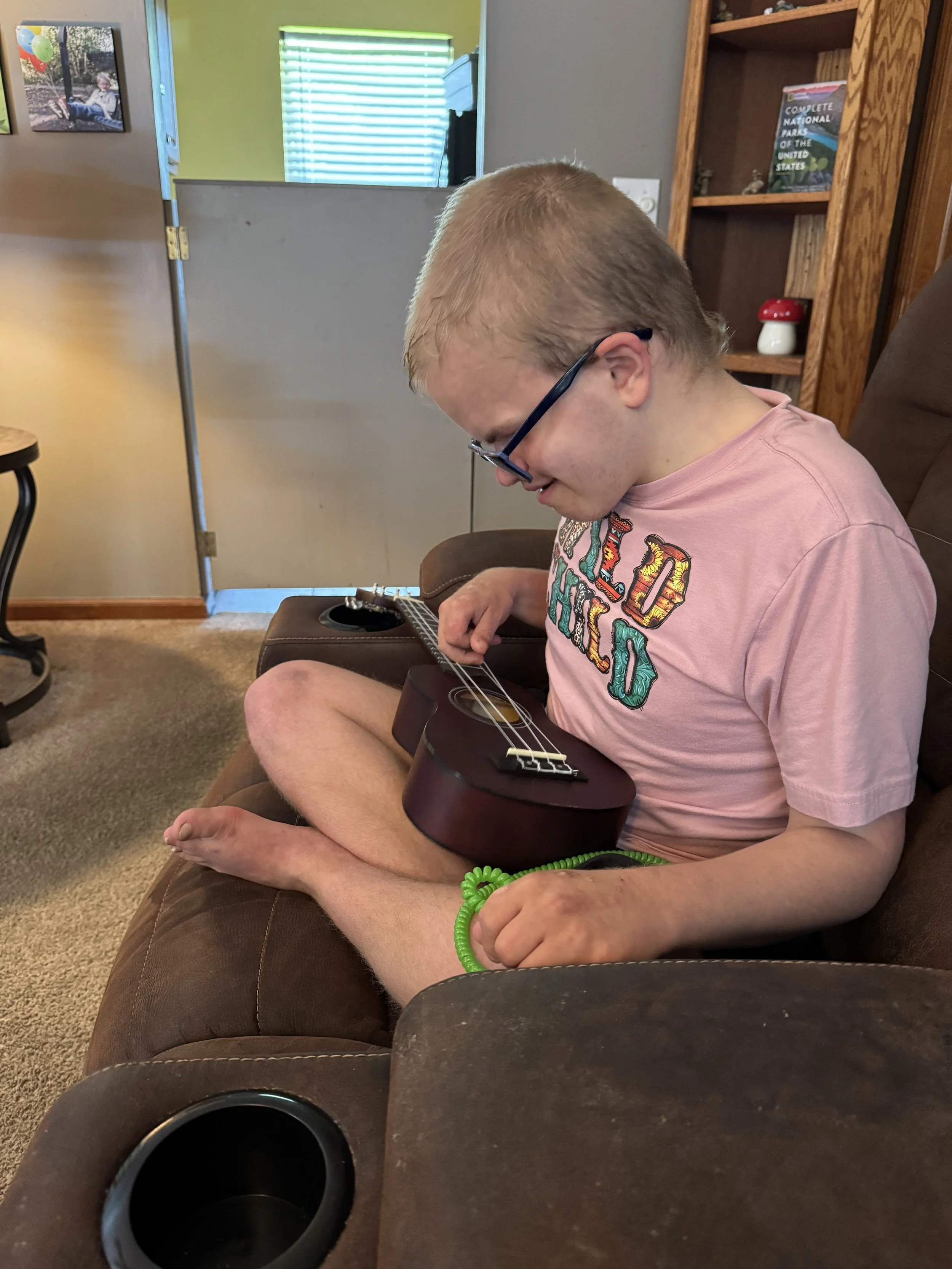 A young boy sitting cross-legged on a couch, playing a small acoustic guitar. He is wearing glasses, a pink T-shirt, and a green bracelet. This is during his music therapy session.