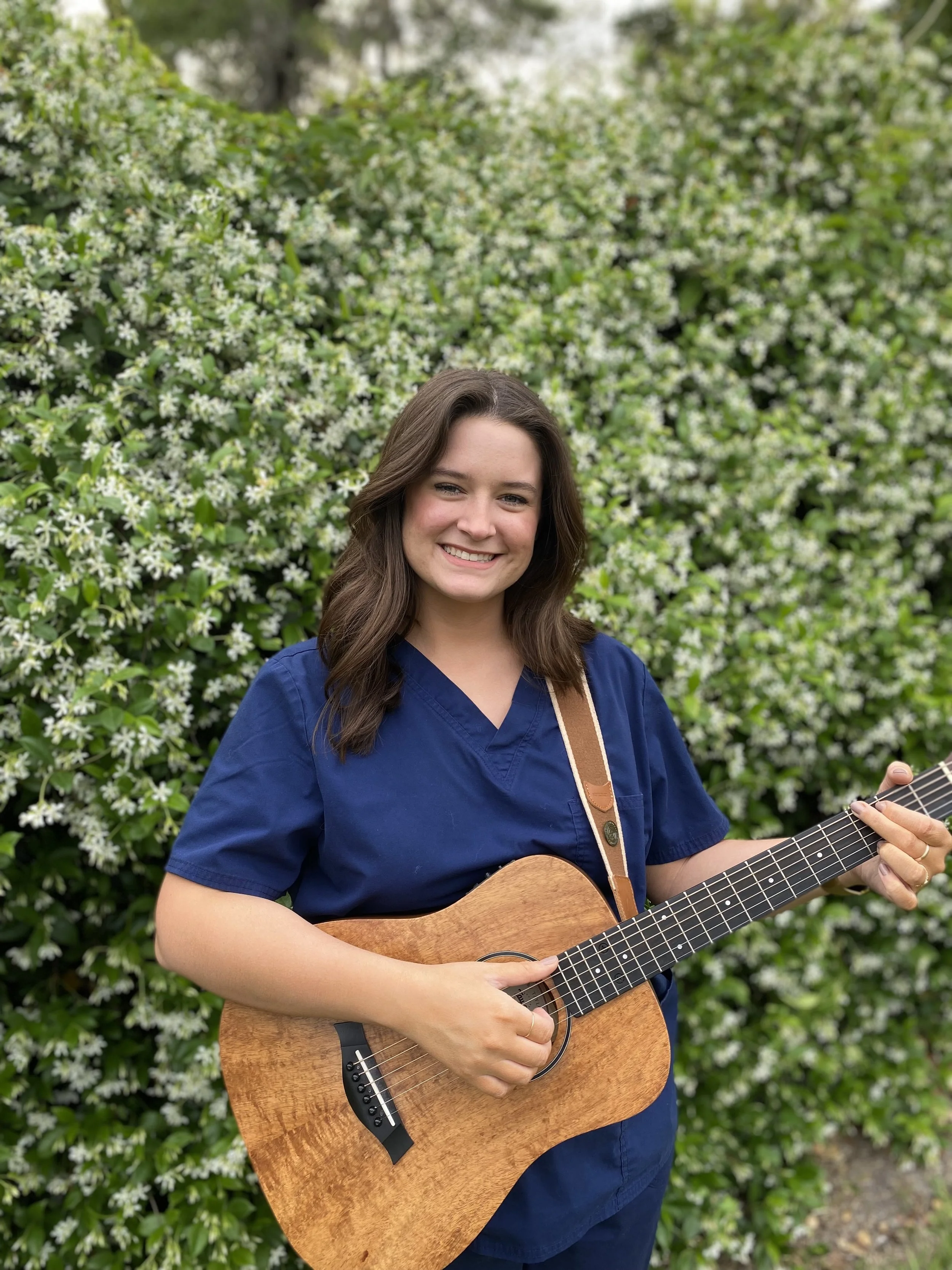 A young woman who is a music therapist in blue medical scrubs playing an acoustic guitar outdoors, standing in front of a bush with white flowers, smiling at the camera.