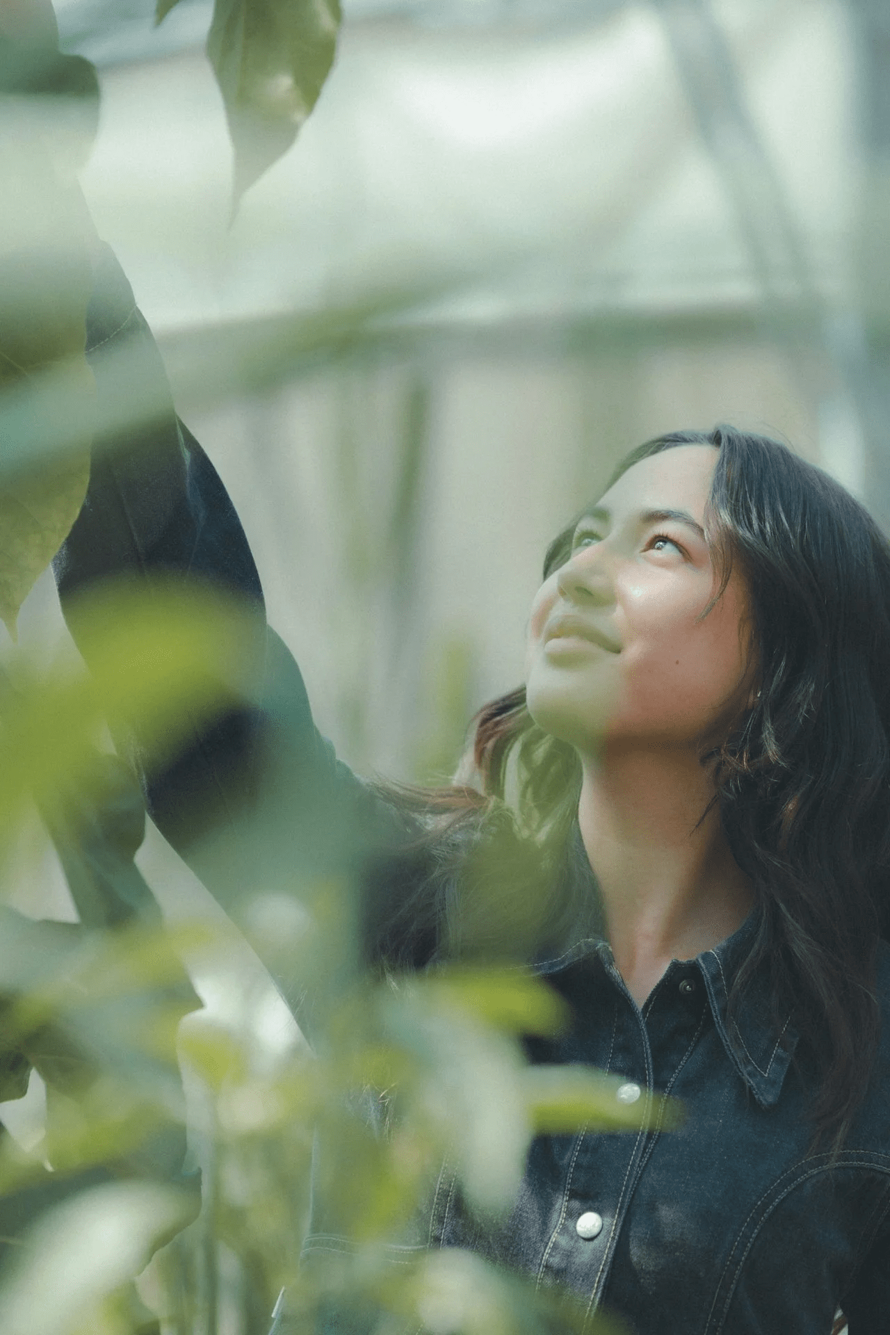 A woman with long dark hair looking up at a person standing beside her in a green outdoor setting, with leaves partially obscuring the foreground.