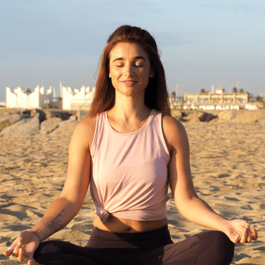 Woman meditating on a sandy beach with white buildings in the background, sitting cross-legged with closed eyes and a peaceful expression.