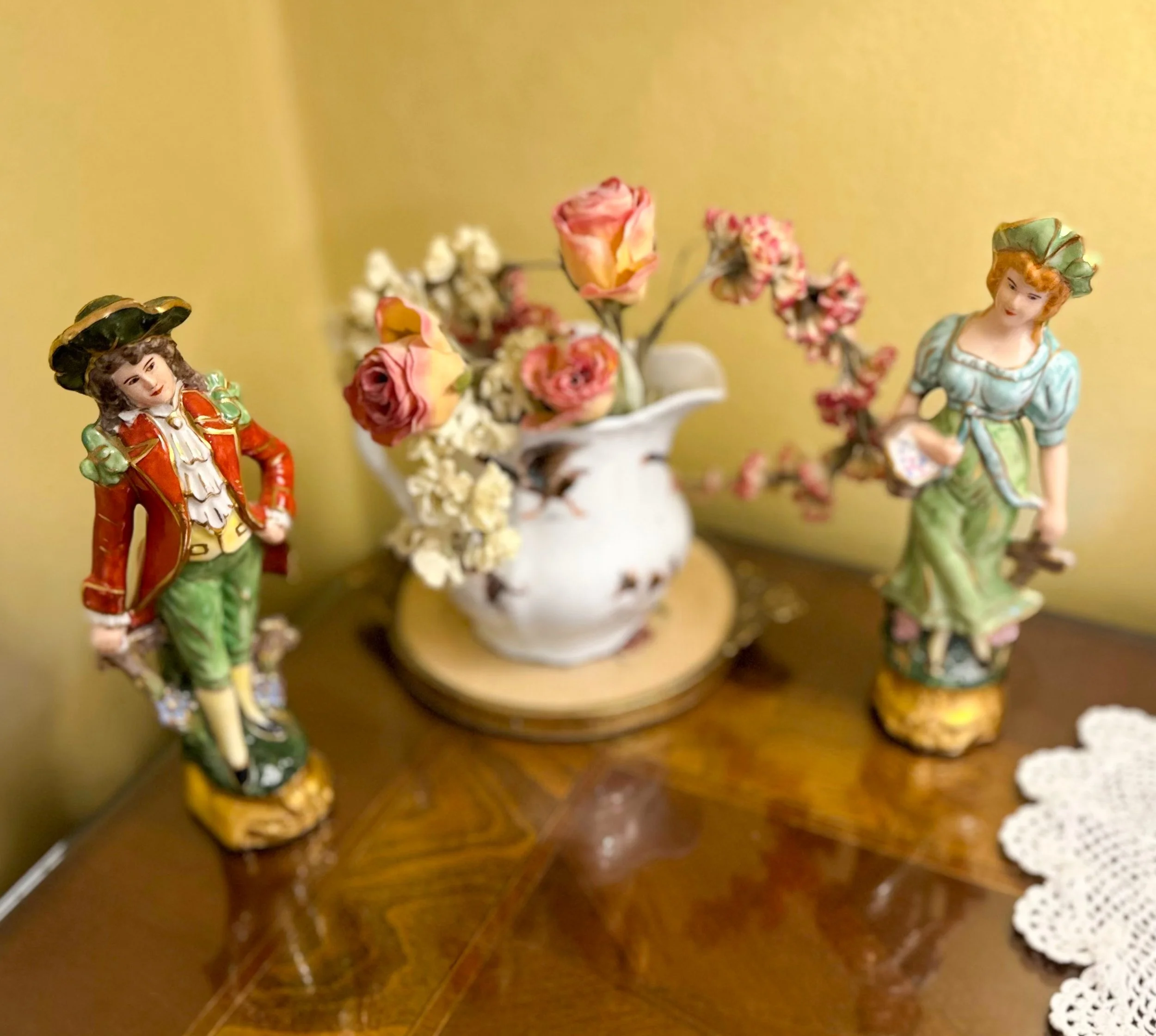 Decorative porcelain figurines of a man and woman beside a ceramic vase with artificial flowers on a wooden table with a lace doily.