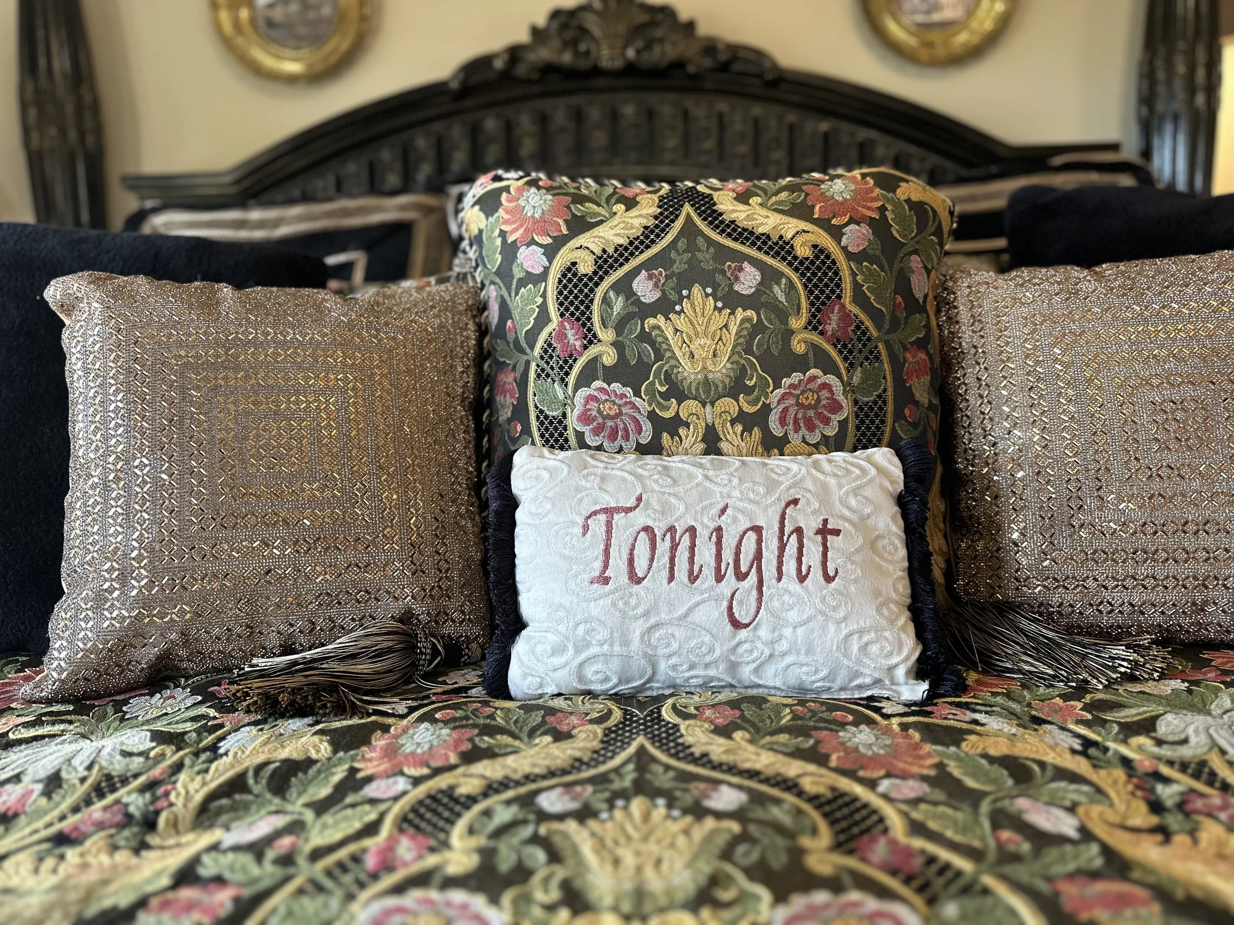 A decorative bed setting with ornate pillows, including one embroidered with "Tonight." The pillows feature intricate patterns and gold detailing, set against a floral patterned bedspread.