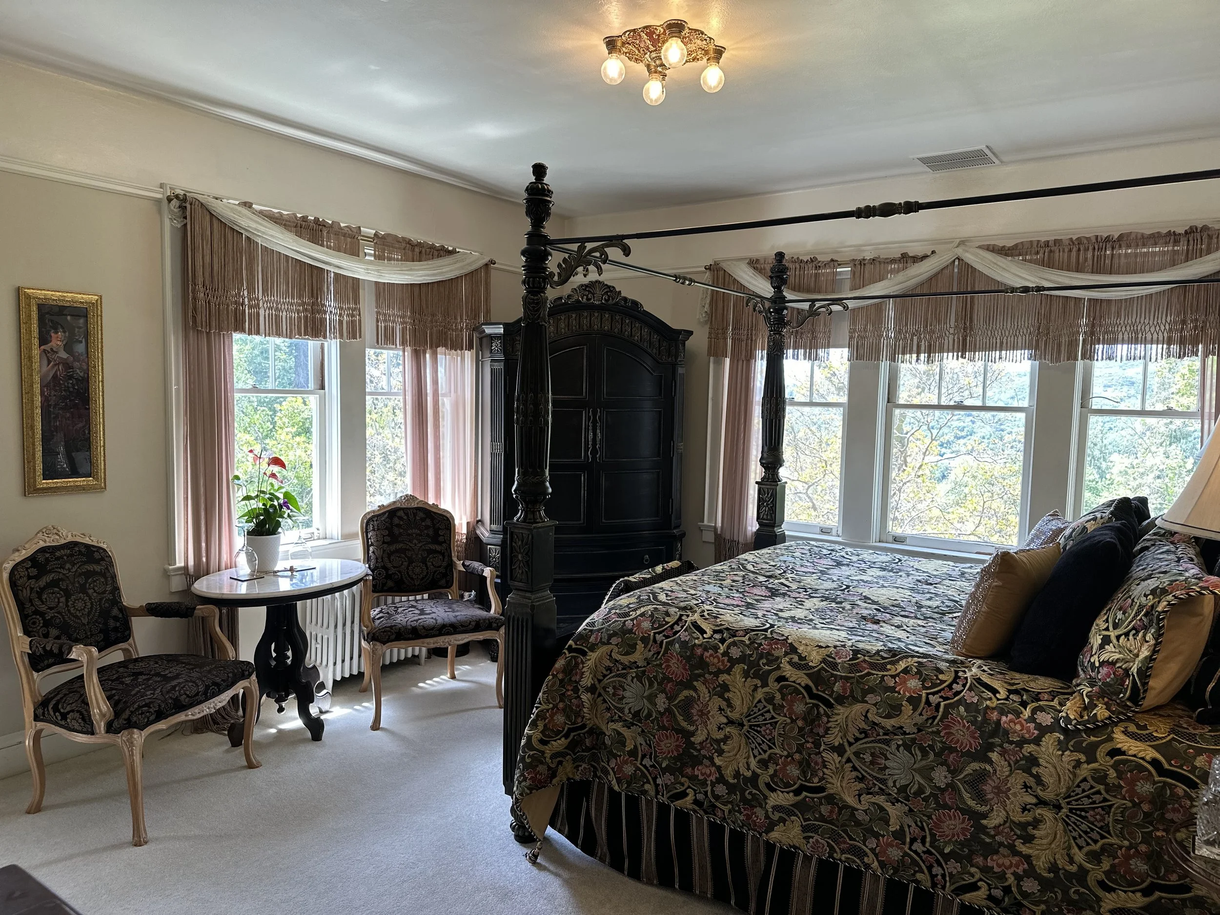 Elegant bedroom with ornate four-poster bed, floral bedding, vintage furniture, two armchairs around a small table, large windows with draped curtains, and a framed wall art.