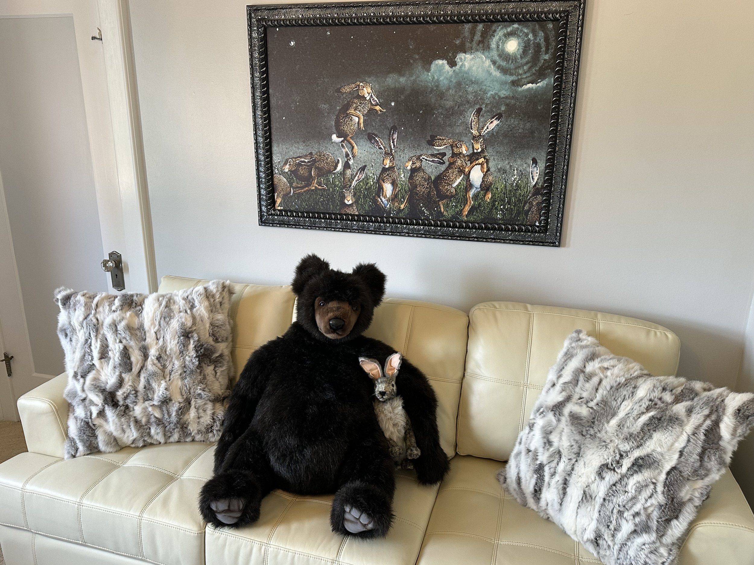 A living room with a cream-colored sofa featuring two gray faux fur pillows. A large teddy bear and a smaller stuffed animal sit on the sofa. Above the sofa hangs a framed painting of rabbits under a moonlit sky.