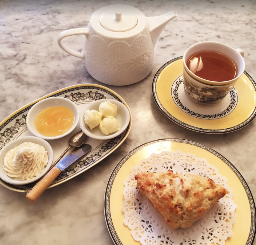 Tea set with a white teapot, teacup on a saucer filled with tea, a scone on a doily-covered plate, and a tray with lemon curd, clotted cream, and butter.