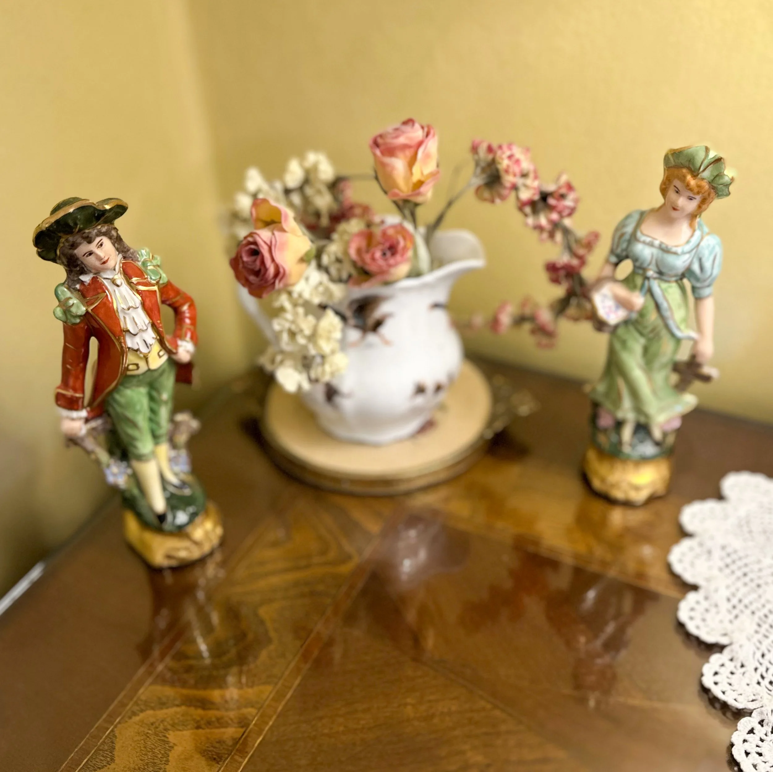 Decorative figurines of a man and woman in vintage clothing, a white vase with pink and white flowers, and a lace doily on a wooden table.