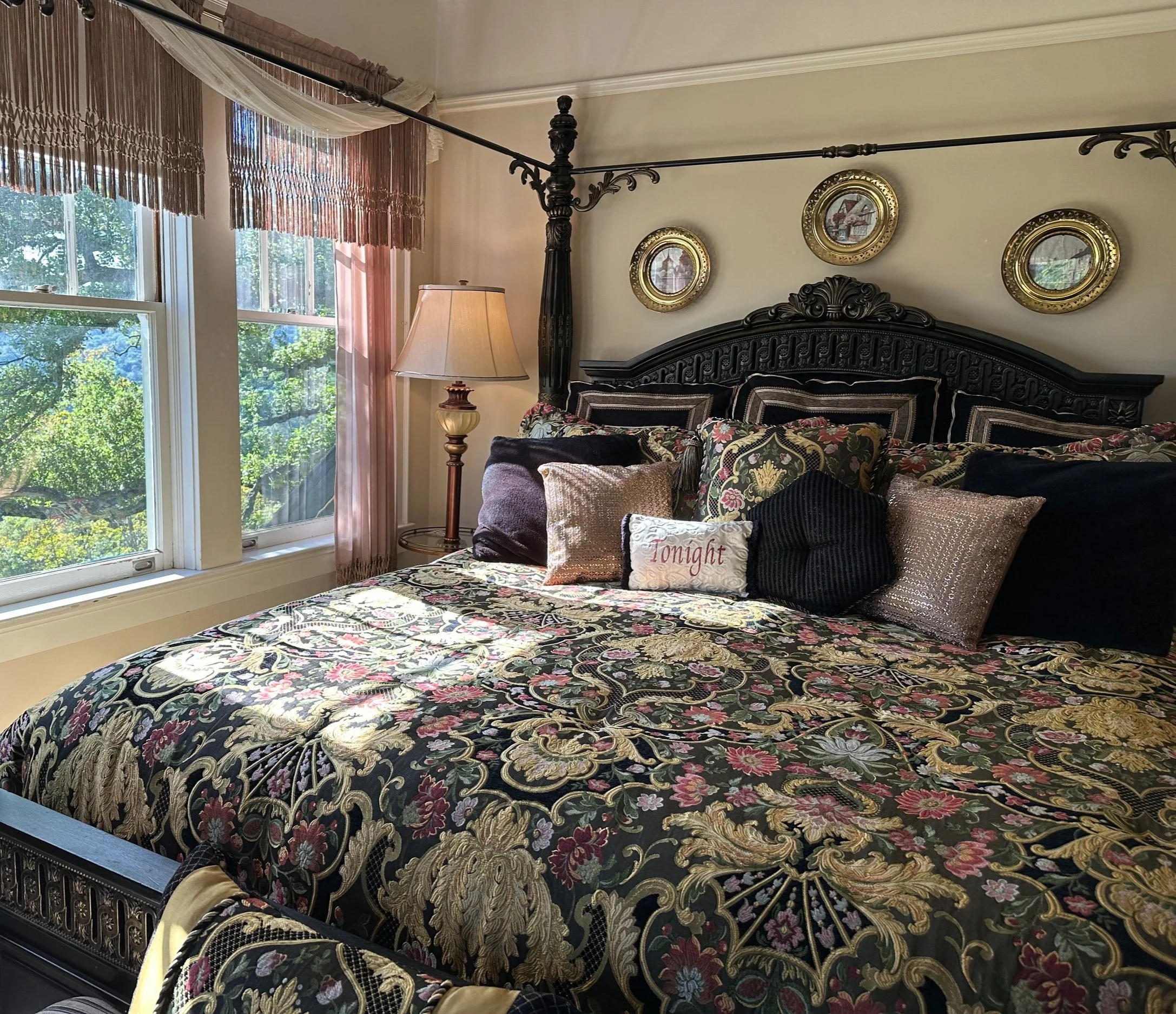 Elegant bedroom with a large bed featuring floral patterned bedding, decorative pillows, and an ornate headboard. The room has a lamp, framed pictures on the wall, and large windows with sheer curtains.