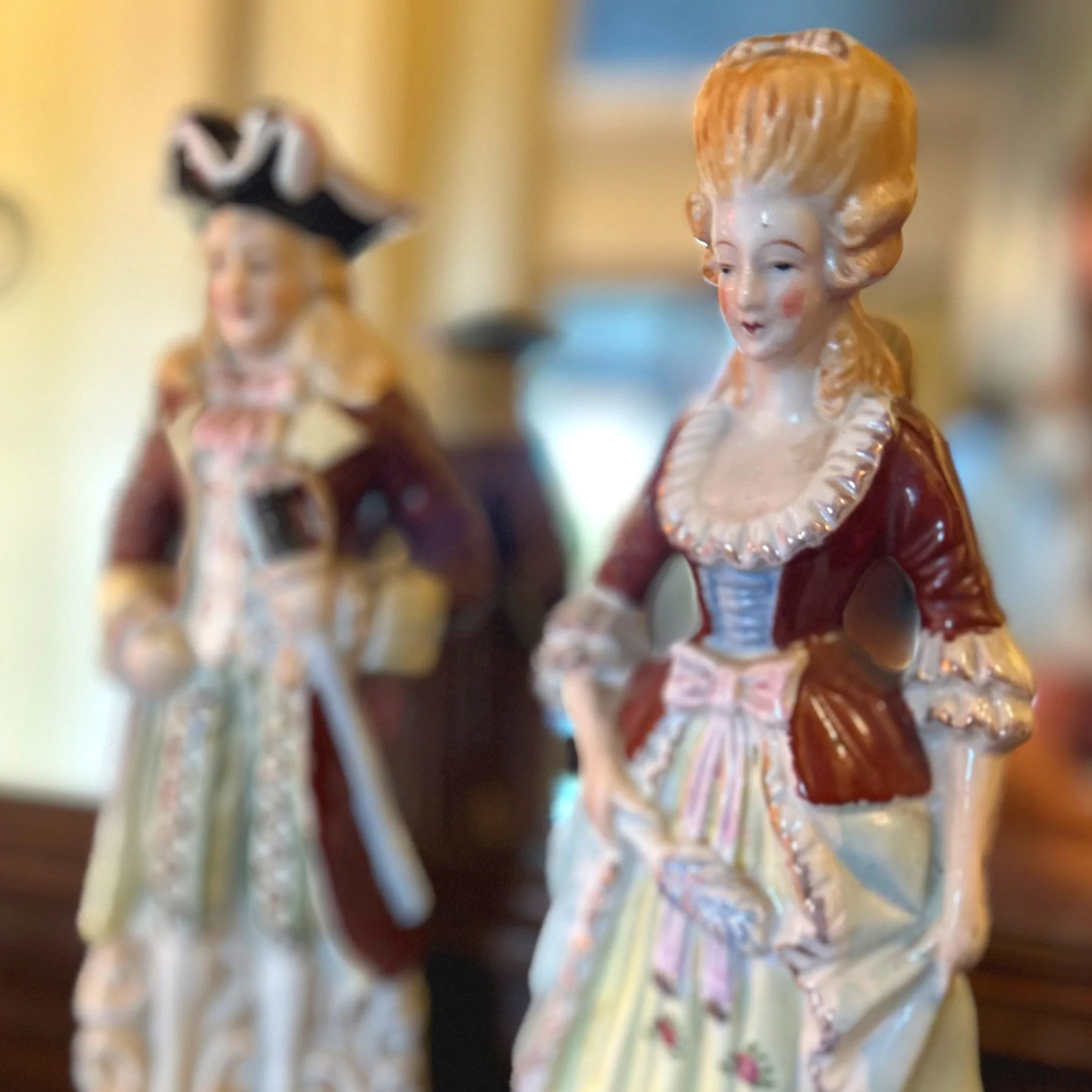 Porcelain figurines of a man and woman in 18th-century attire, with the woman in the foreground wearing a gown and holding a fan, and the man in the background wearing a tricorn hat and coat, both with detailed clothing and expressions.