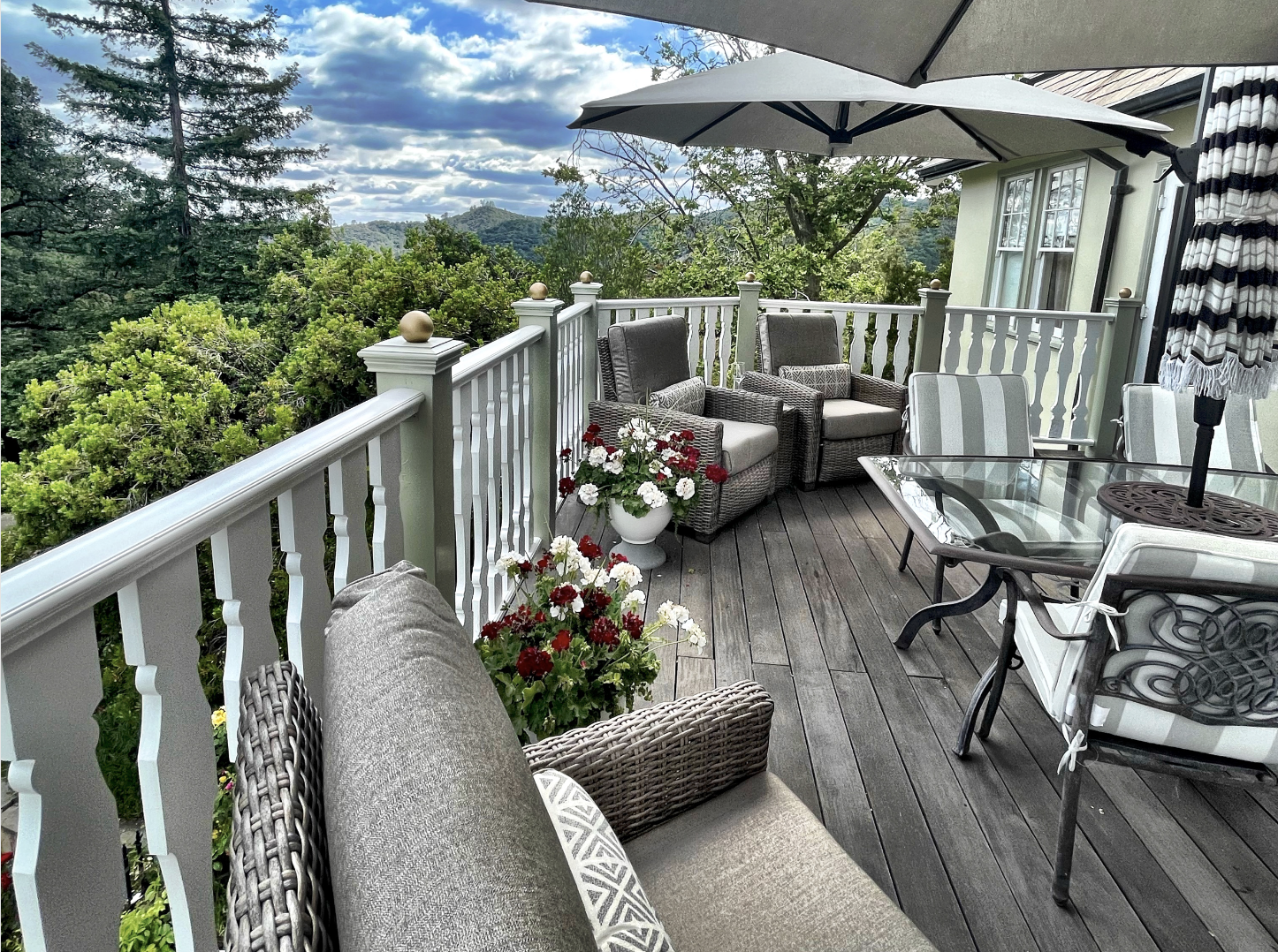 Scenic patio view with wicker furniture, striped cushions, glass table, and umbrella on a wooden deck surrounded by greenery and mountain landscape.