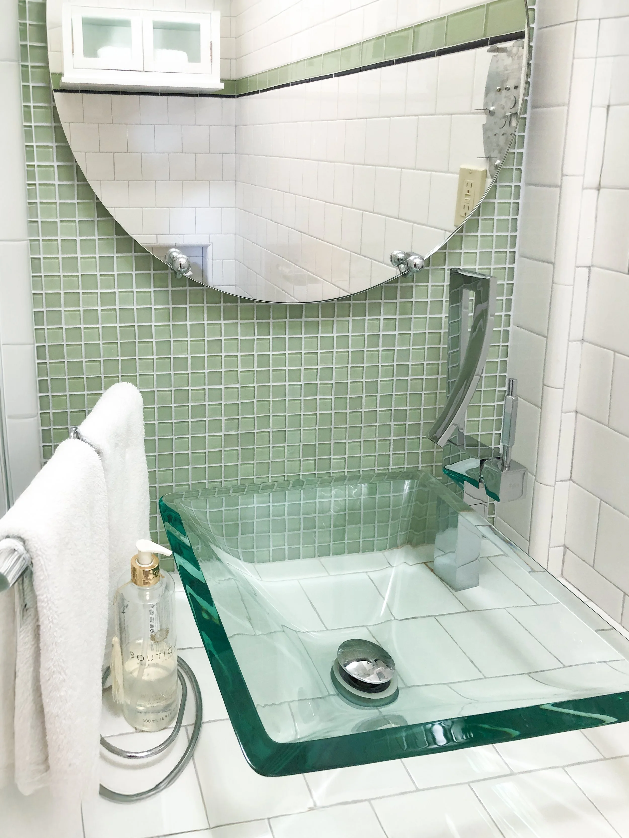 Bathroom with green tiled walls, a glass sink with a silver drain, a round mirror reflecting tiled walls and a window, a towel rack with white towels, a soap dispenser, and a modern faucet.