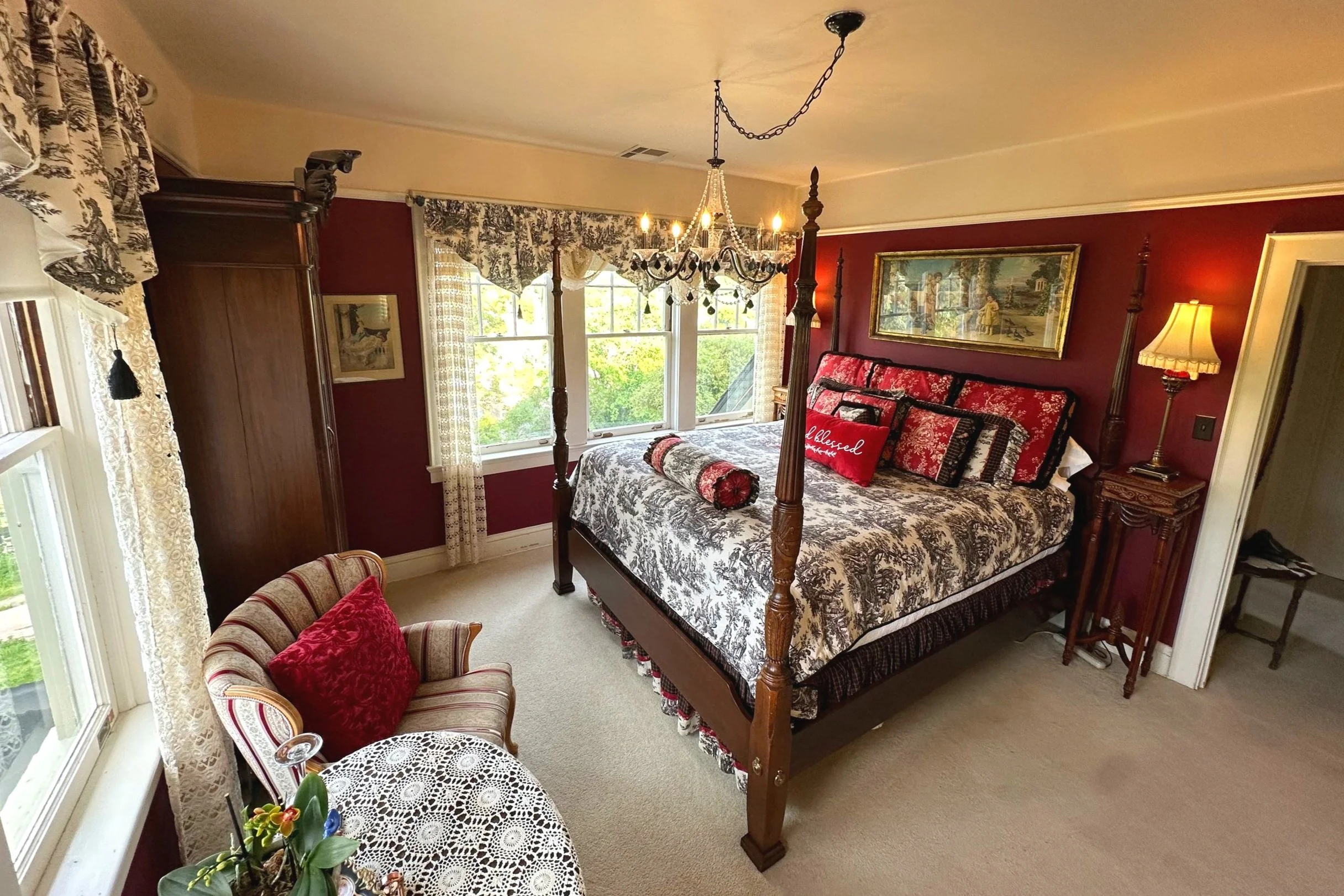 Elegant bedroom with a vintage four-poster bed, ornate chandelier, and red decor. Features a striped armchair with a red pillow, lace curtains, and a small round table with a crochet tablecloth. The room has rich red walls and is decorated with framed art and a standing lamp.