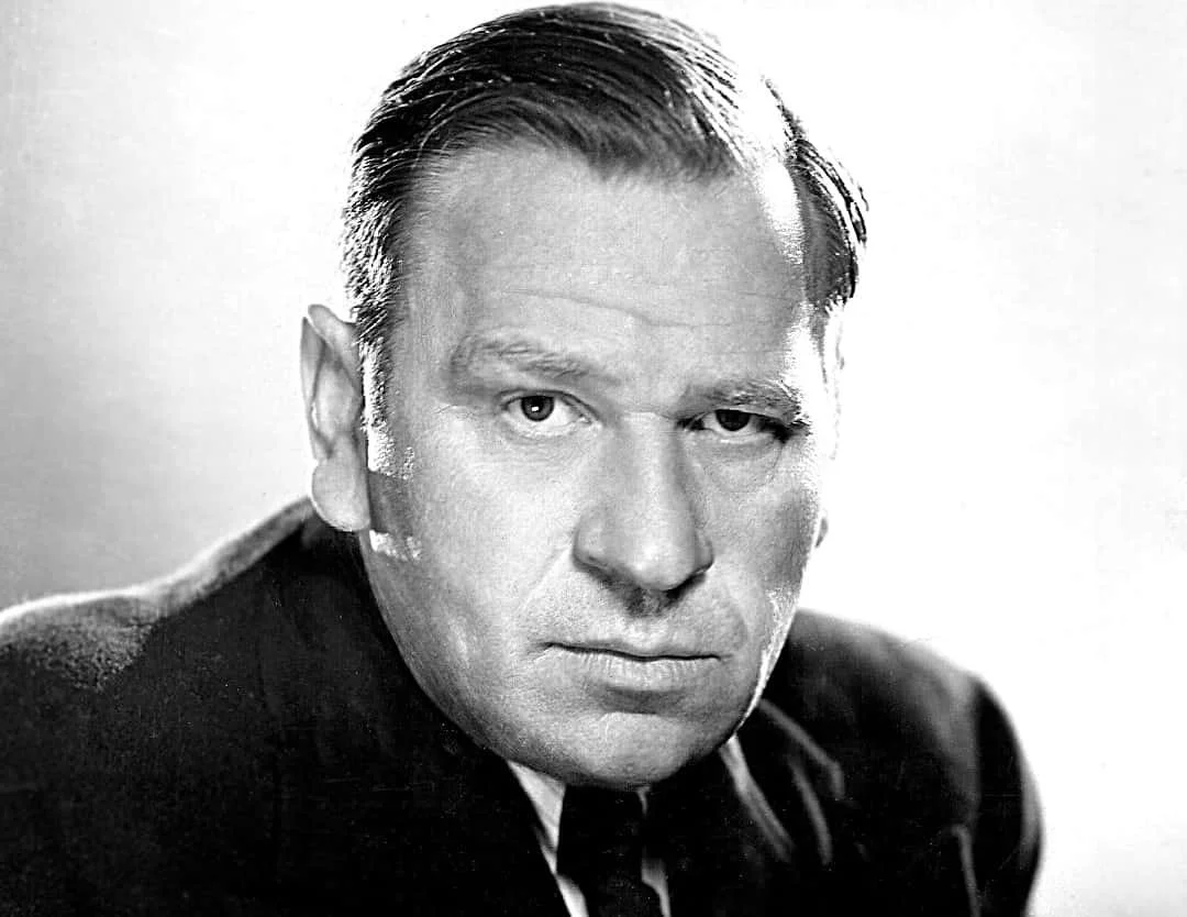 Black and white portrait of a man with slicked-back hair, wearing a dark top, with serious expression