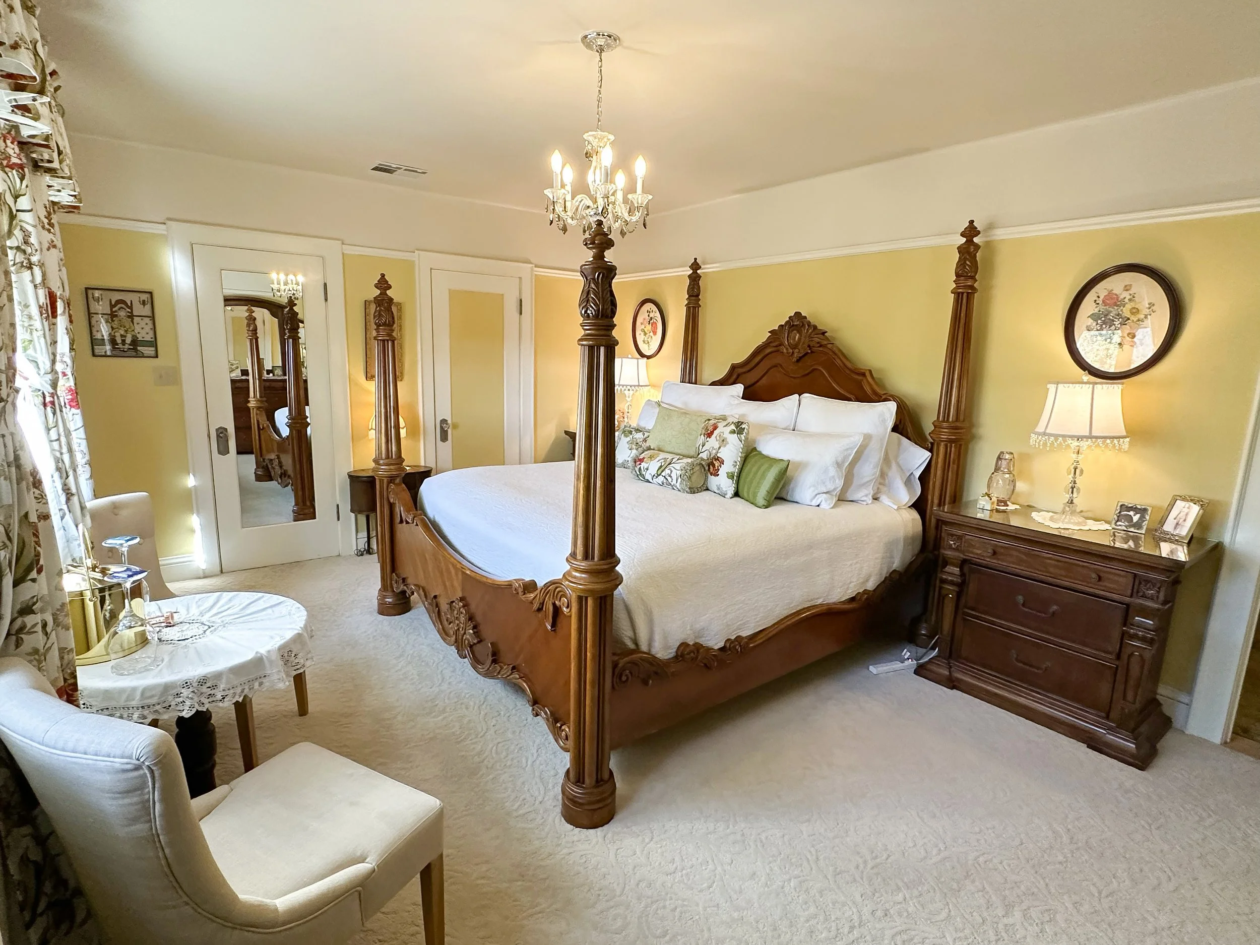 A bedroom with a large wooden four-poster bed, side table with lamp and framed photographs, cream-colored armchair, small round table with glasses and a wine bottle, floral curtains, yellow walls, and double doors with mirrors.