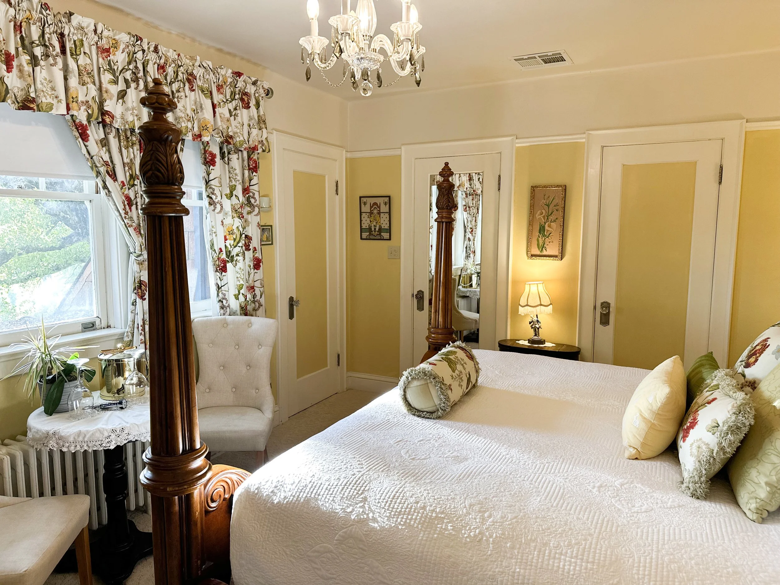 A cozy bedroom with a large four-poster wooden bed, floral pillows, a bedside lamp, a window with floral curtains, a white upholstered chair, and decorative artwork. The room has soft yellow walls and a chandelier.