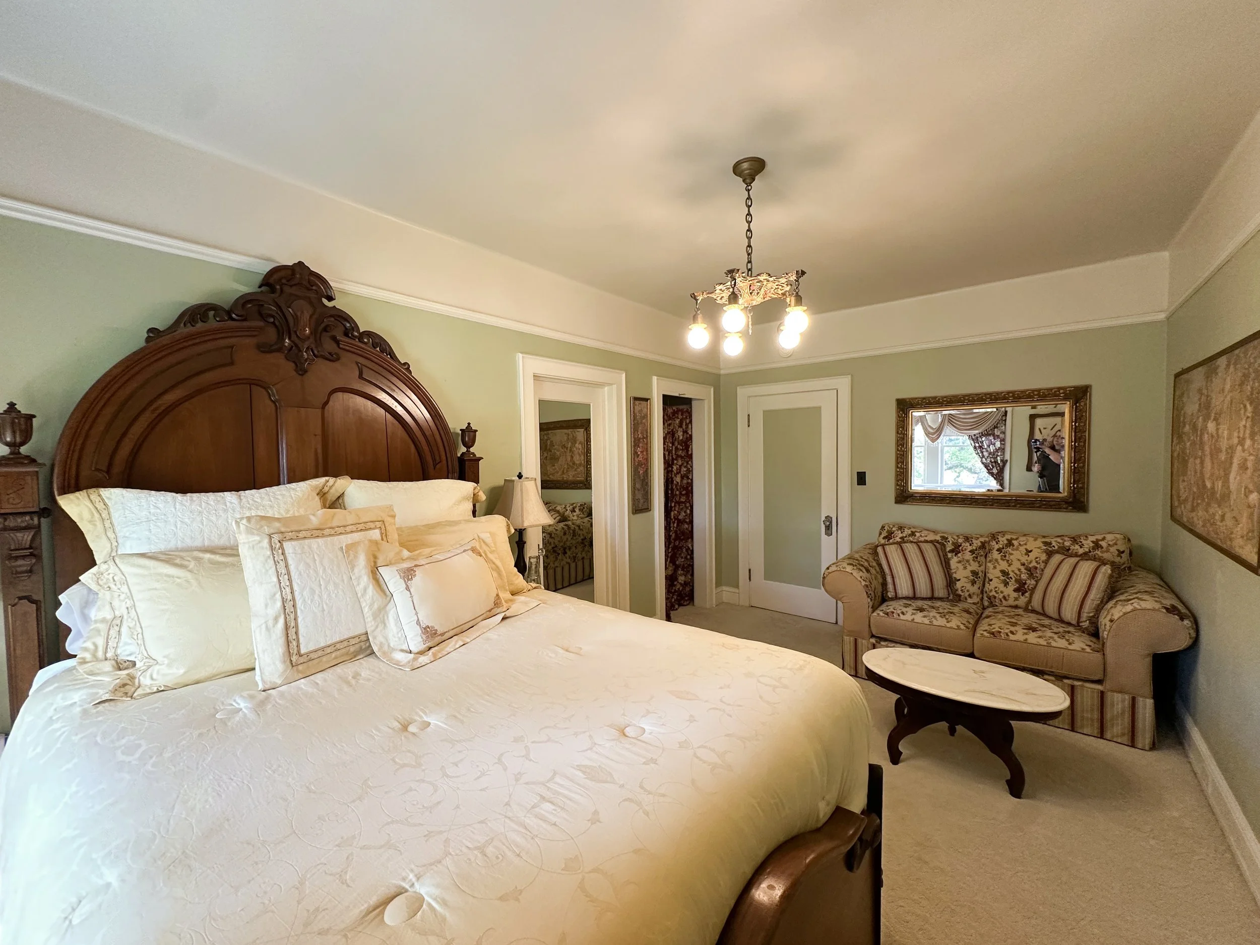 A bedroom with a large wooden bed with multiple pillows, a side table with a lamp, a floral sofa with cushions, a small coffee table, framed mirror on the wall, and a chandelier hanging from the ceiling.