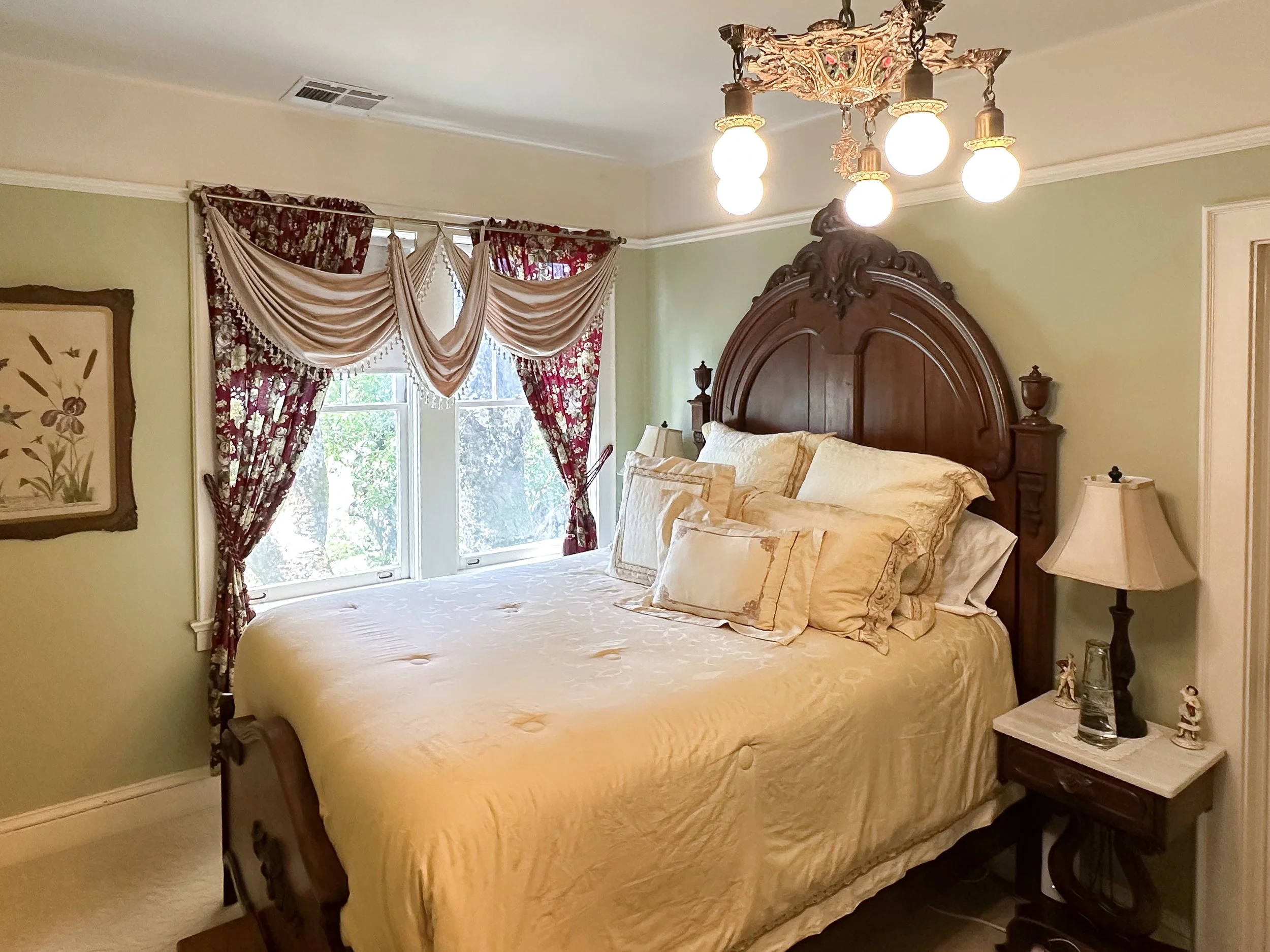 A bedroom with green walls, a large wooden headboard, cream-colored bedding, a nightstand with a lamp, and a window with floral and beige curtains.