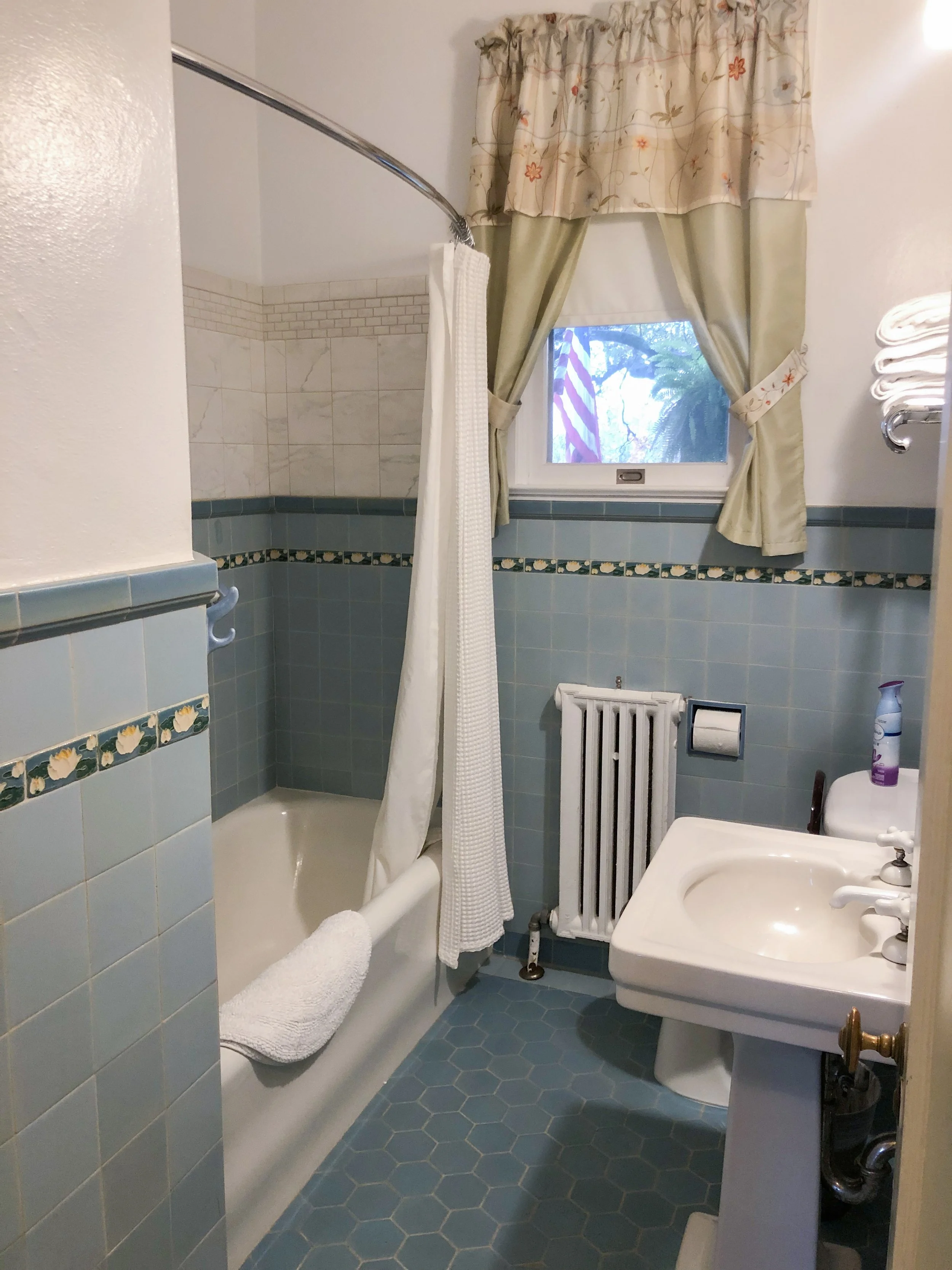 Small bathroom with a shower, window with curtains, radiator, and sink, all in a blue and white color scheme.