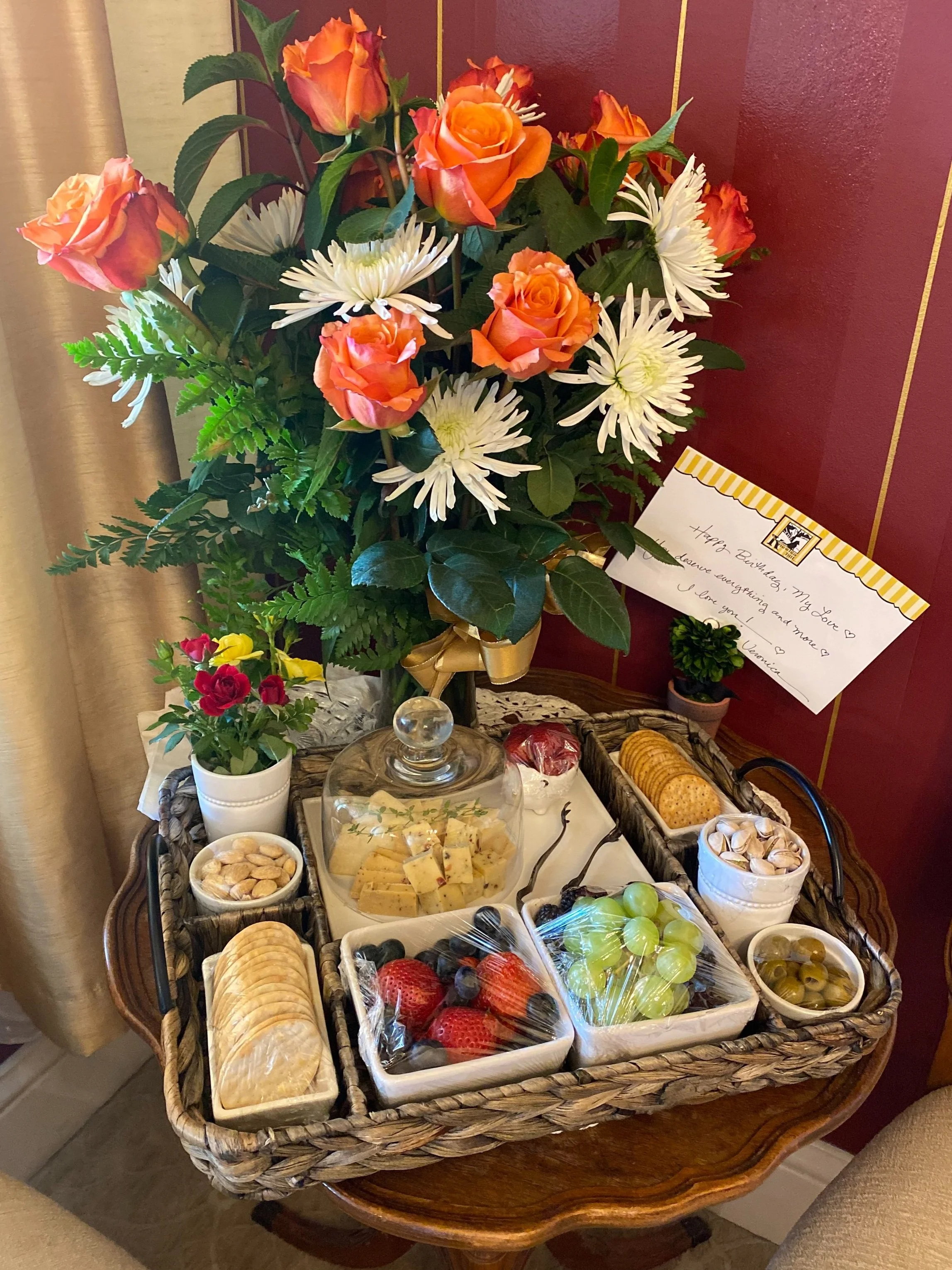 A wicker tray on a table contains a variety of snacks including sliced cheese under a glass dome, crackers, mixed nuts, olives, strawberries, grapes, and other fruits. Beside the tray is a bouquet of orange roses and white flowers, and a card with a handwritten message.