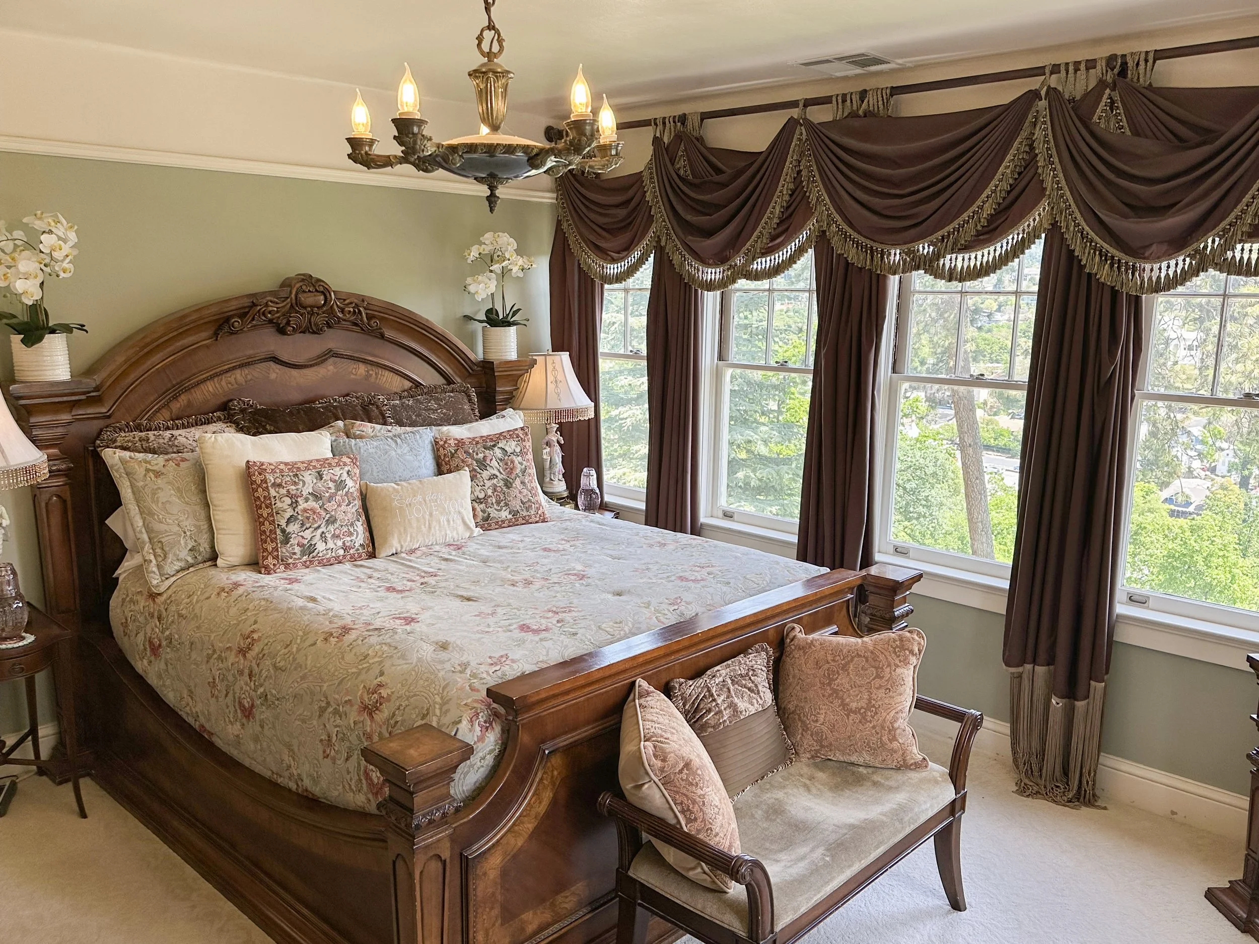 Elegant bedroom with ornate wooden bed, floral bedding, and multiple decorative pillows. Large windows with brown drapes overlook greenery. A chandelier hangs above, flanked by lamps and potted orchids.