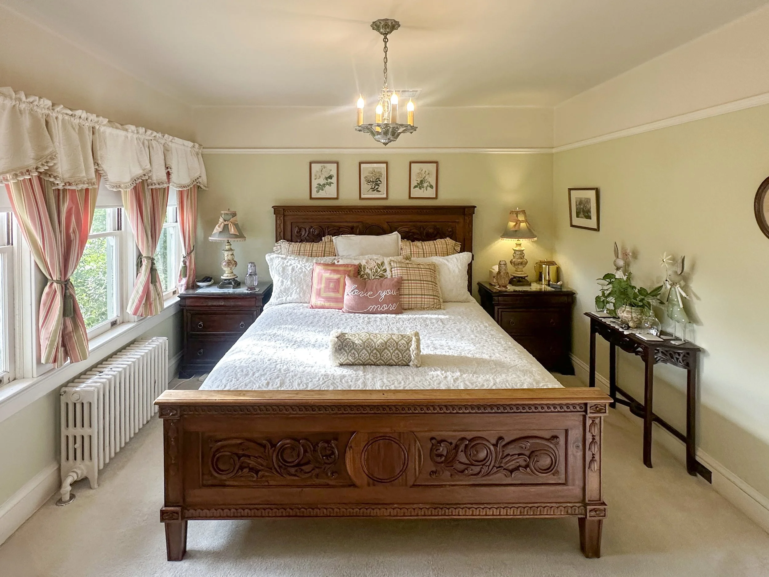 A cozy bedroom with a wooden bed, framed botanical artwork, floral curtains, and bedside lamps, decorated in a traditional style.