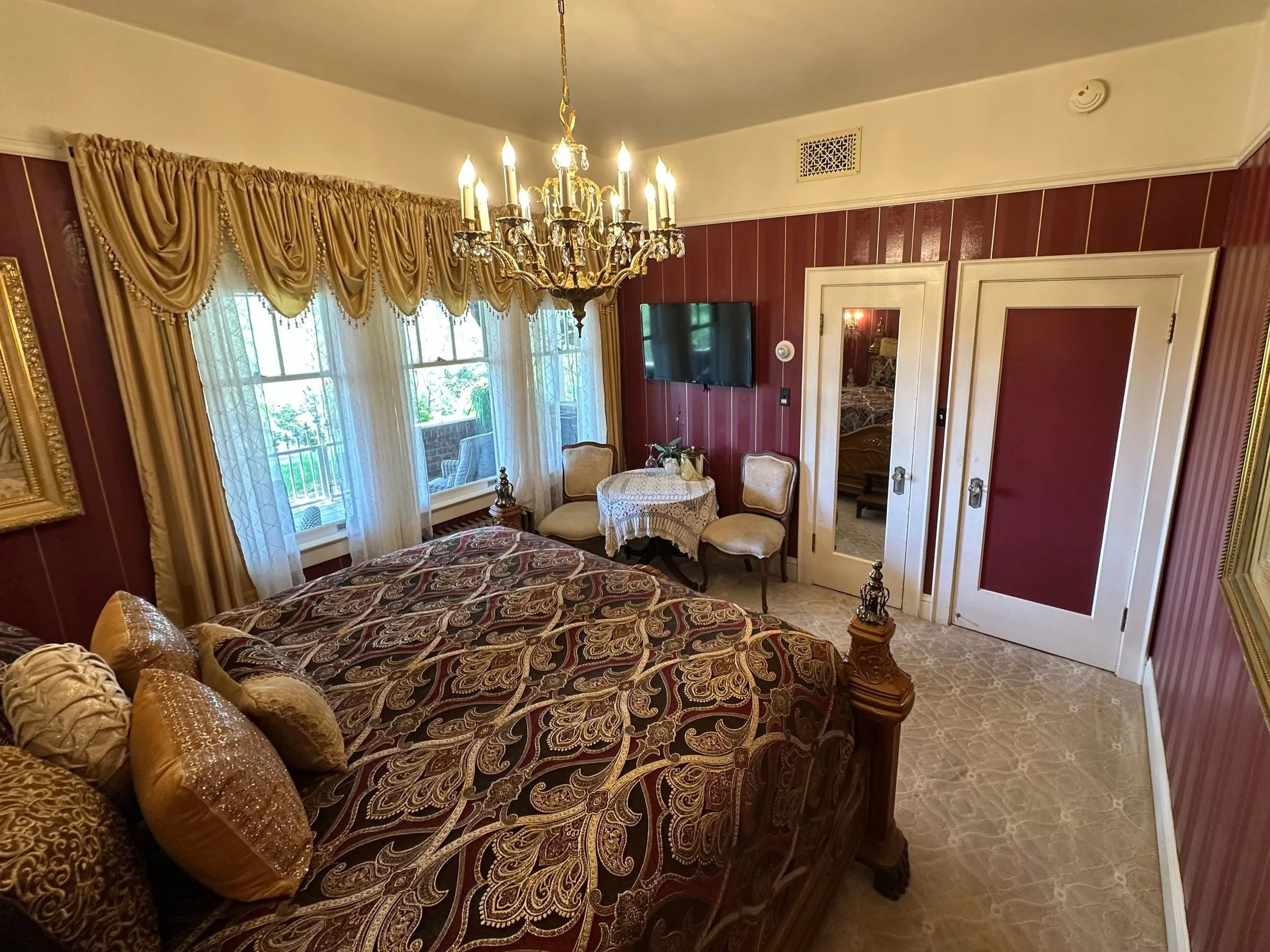 Elegant bedroom interior with a patterned bedspread and decorative pillows, a chandelier, ornate curtains, a small table with chairs, wall-mounted TV, and framed art on walls.