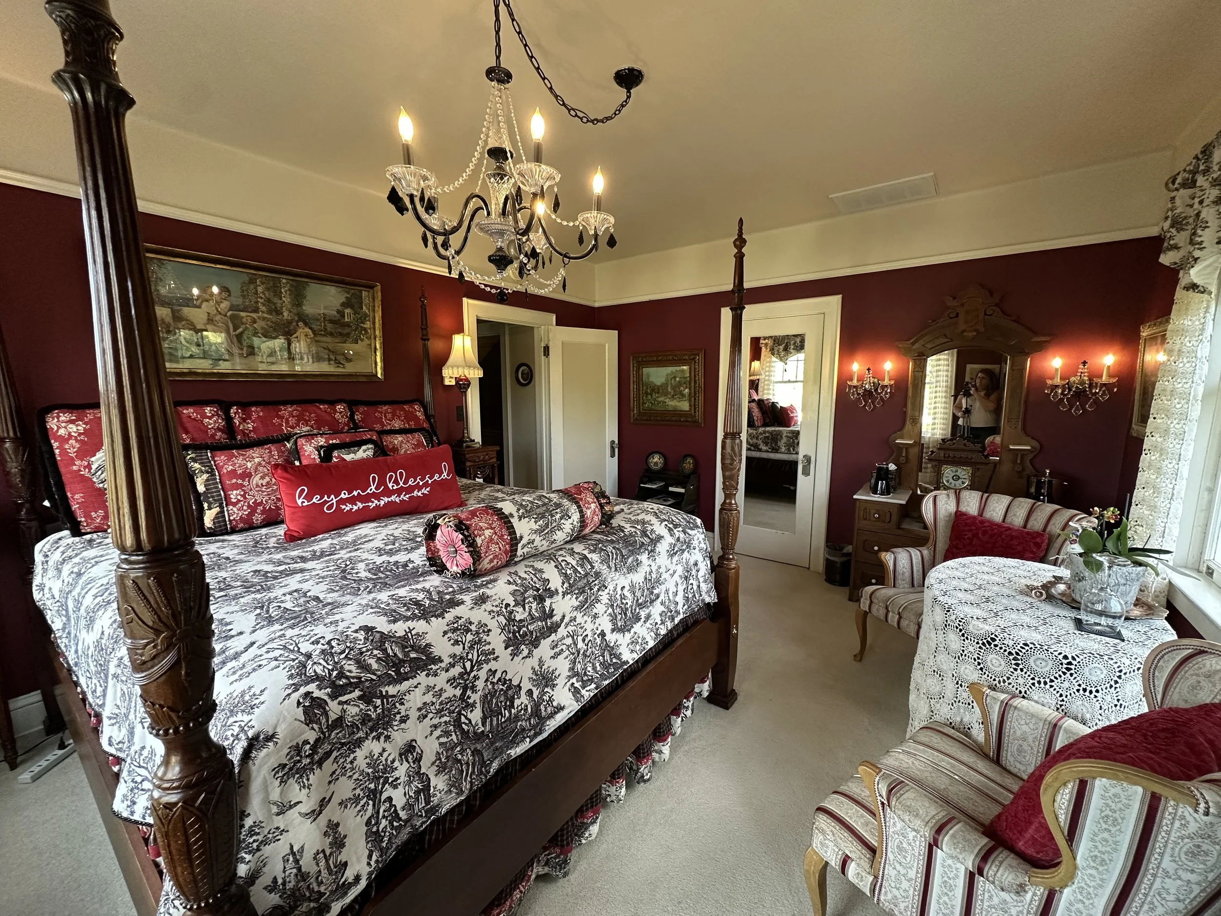 Elegant bedroom with a four-poster bed, ornate chandelier, and vintage-style furniture. The bedding features a toile pattern, and a red pillow reads "beyond blessed." There is a small seating area with striped chairs and a round table with a lace tablecloth.
