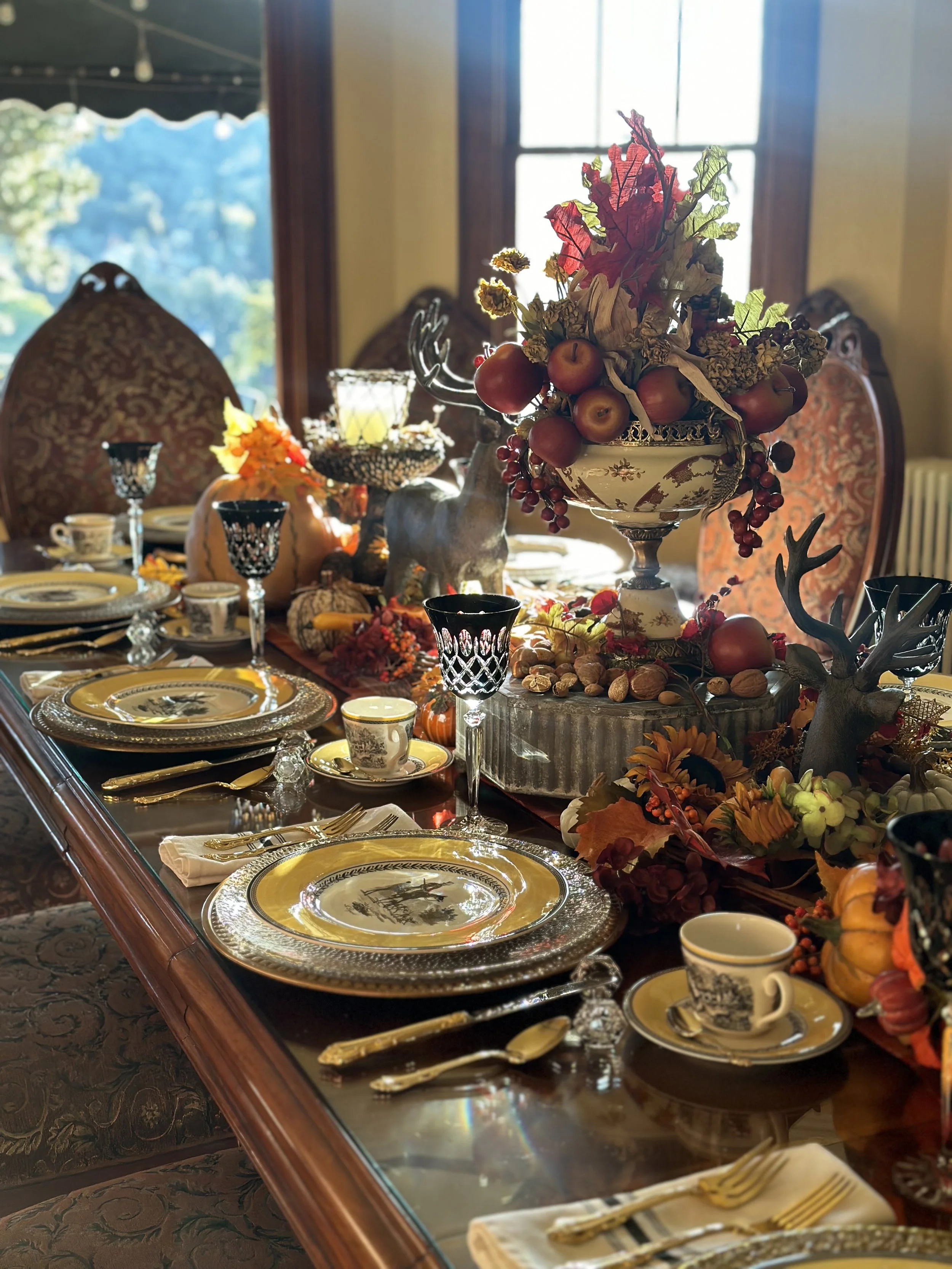 Elegant dining table set with autumn-themed decorations, including a centerpiece of fruit and foliage, ornate dinnerware, glassware, and silverware, ready for a festive meal.