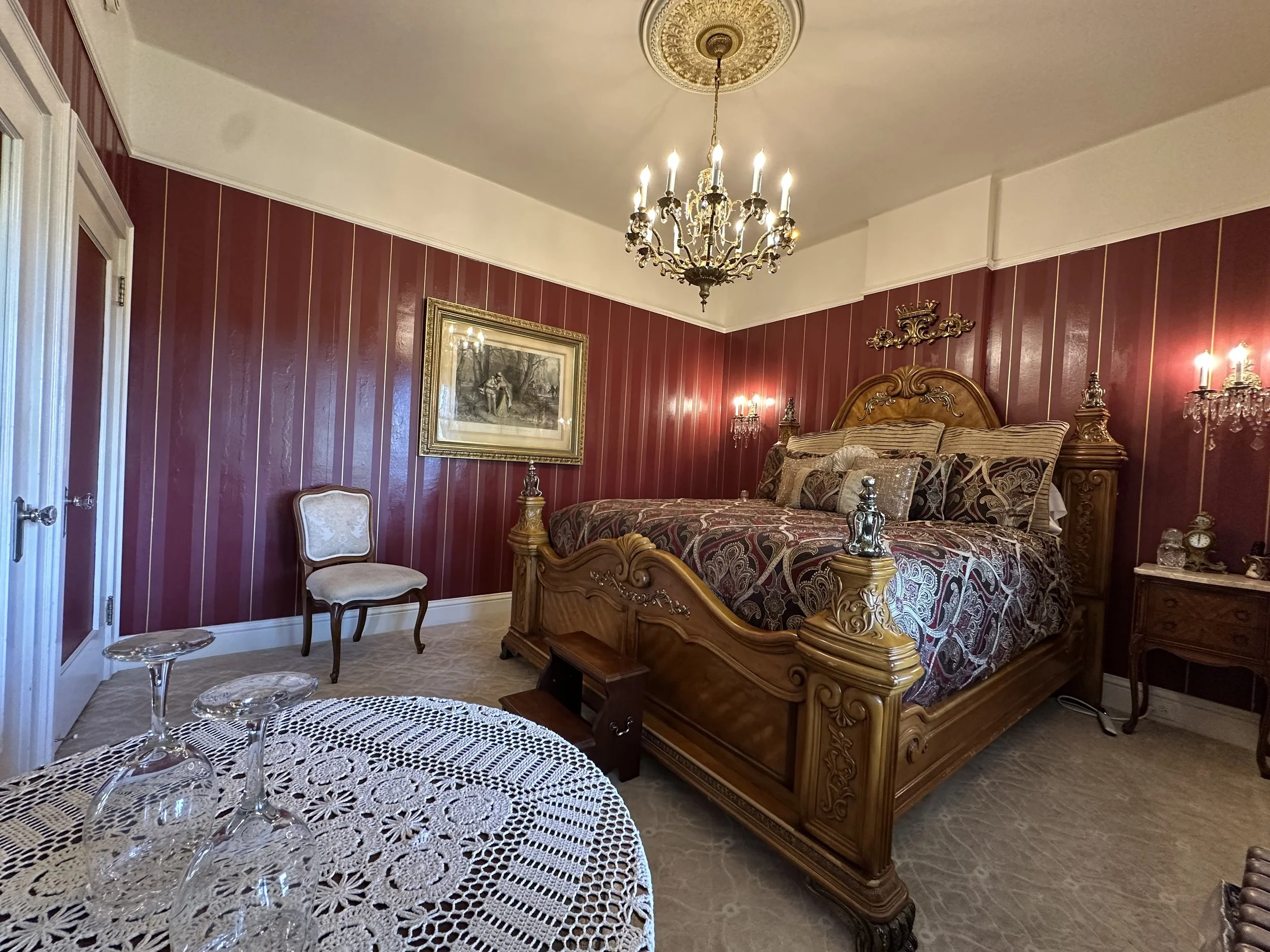 A luxurious bedroom with a wooden bed, ornate headboard, and patterned bedding. The walls are red with vertical gold stripes, and there is a chandelier hanging from the ceiling. The room contains a small side table, a vintage armchair, and lace-covered table with glassware.