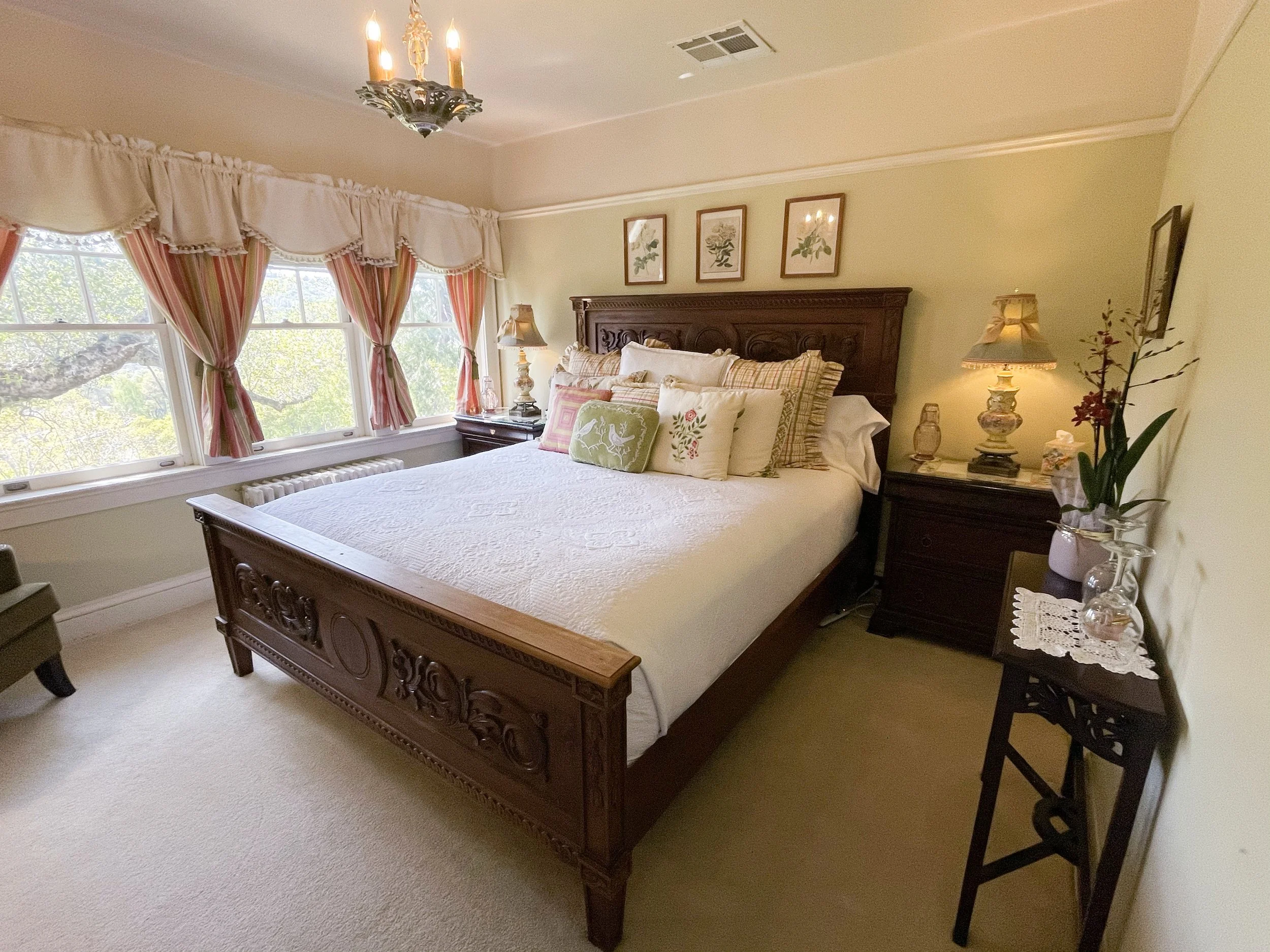 Elegantly decorated bedroom with carved wooden bed, floral pillows, and soft lighting, featuring large windows with lace curtains and a chandelier.