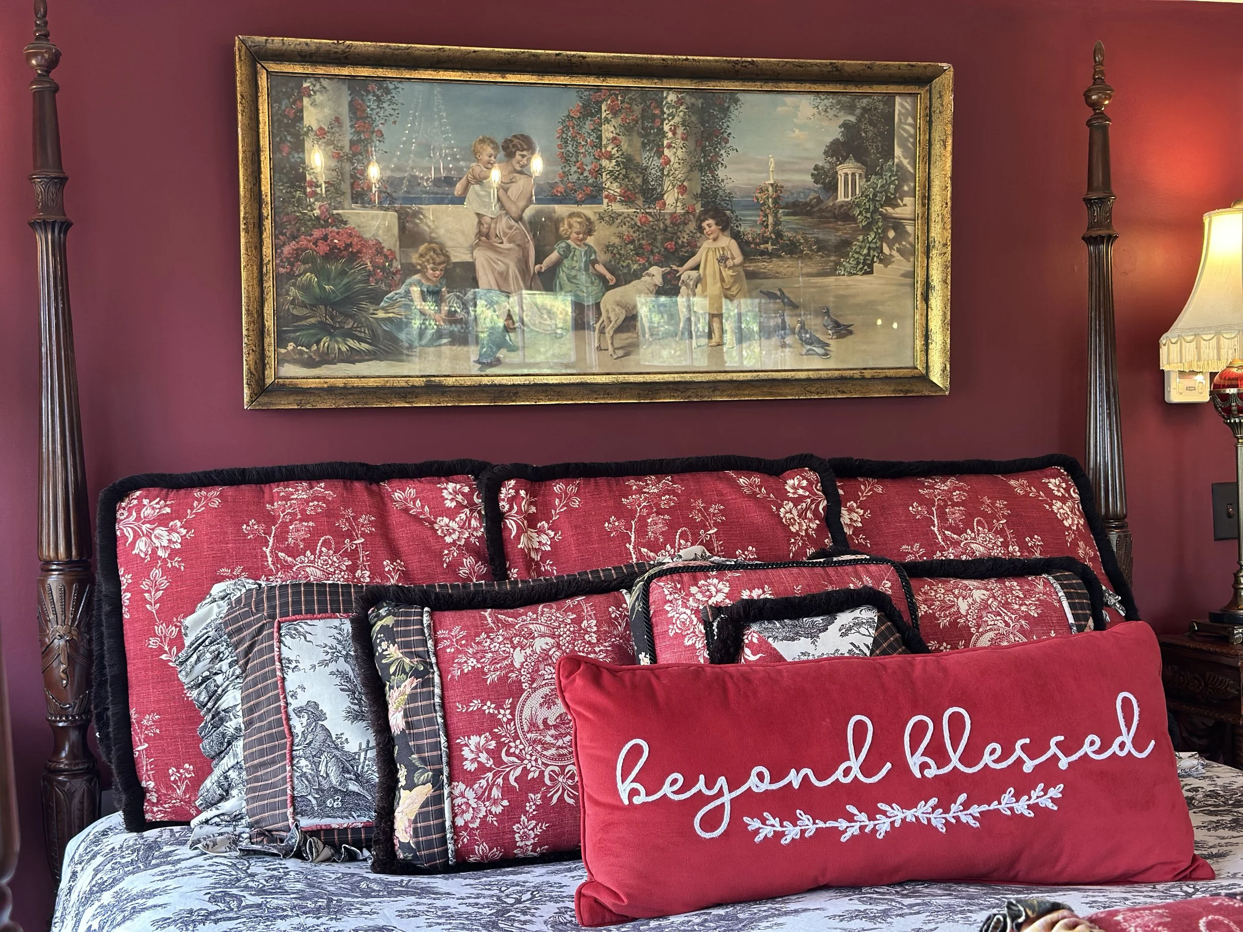 Elegant bedroom with red floral pillows, an embroidered "beyond blessed" pillow, a bed with dark wood posts, and a vintage framed painting on a red wall, depicting children and a garden scene. A lamp is visible on the side.