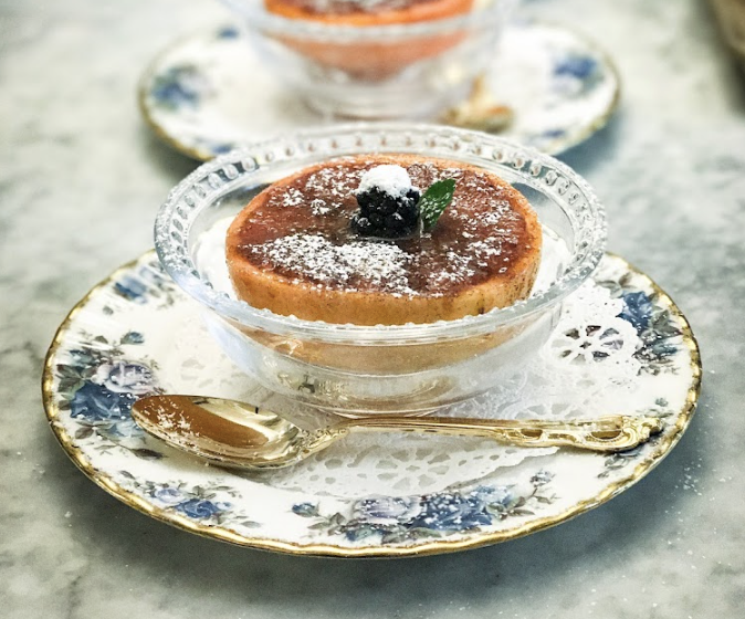 Dessert in glass dish with blackberry, mint, and powdered sugar on floral plate with gold spoon.