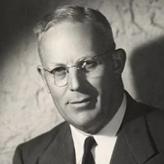 A black and white portrait of a man wearing glasses and a suit, with short hair.