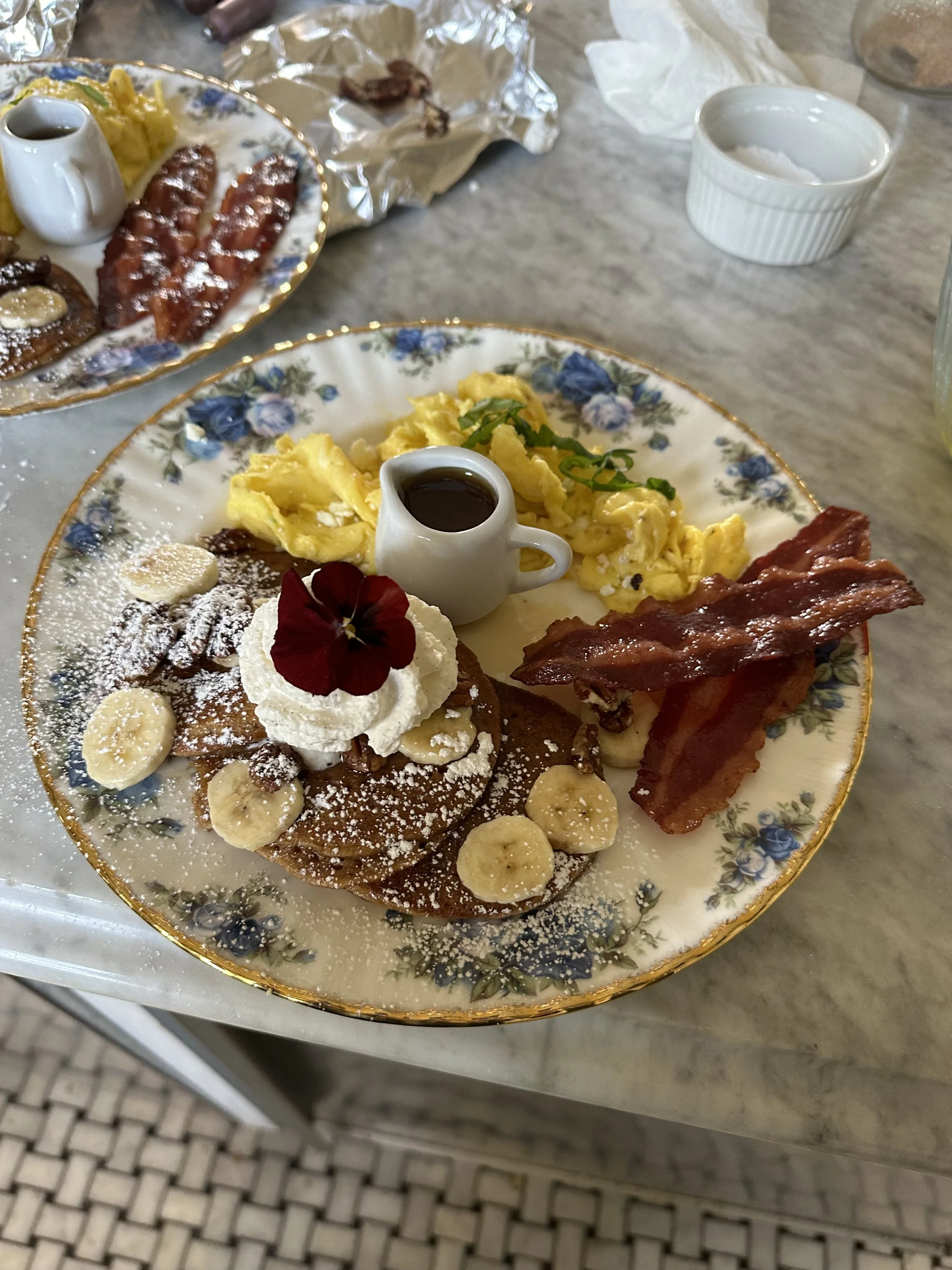 Breakfast plate with banana pancakes, whipped cream, syrup, scrambled eggs, and bacon on a decorative floral plate.