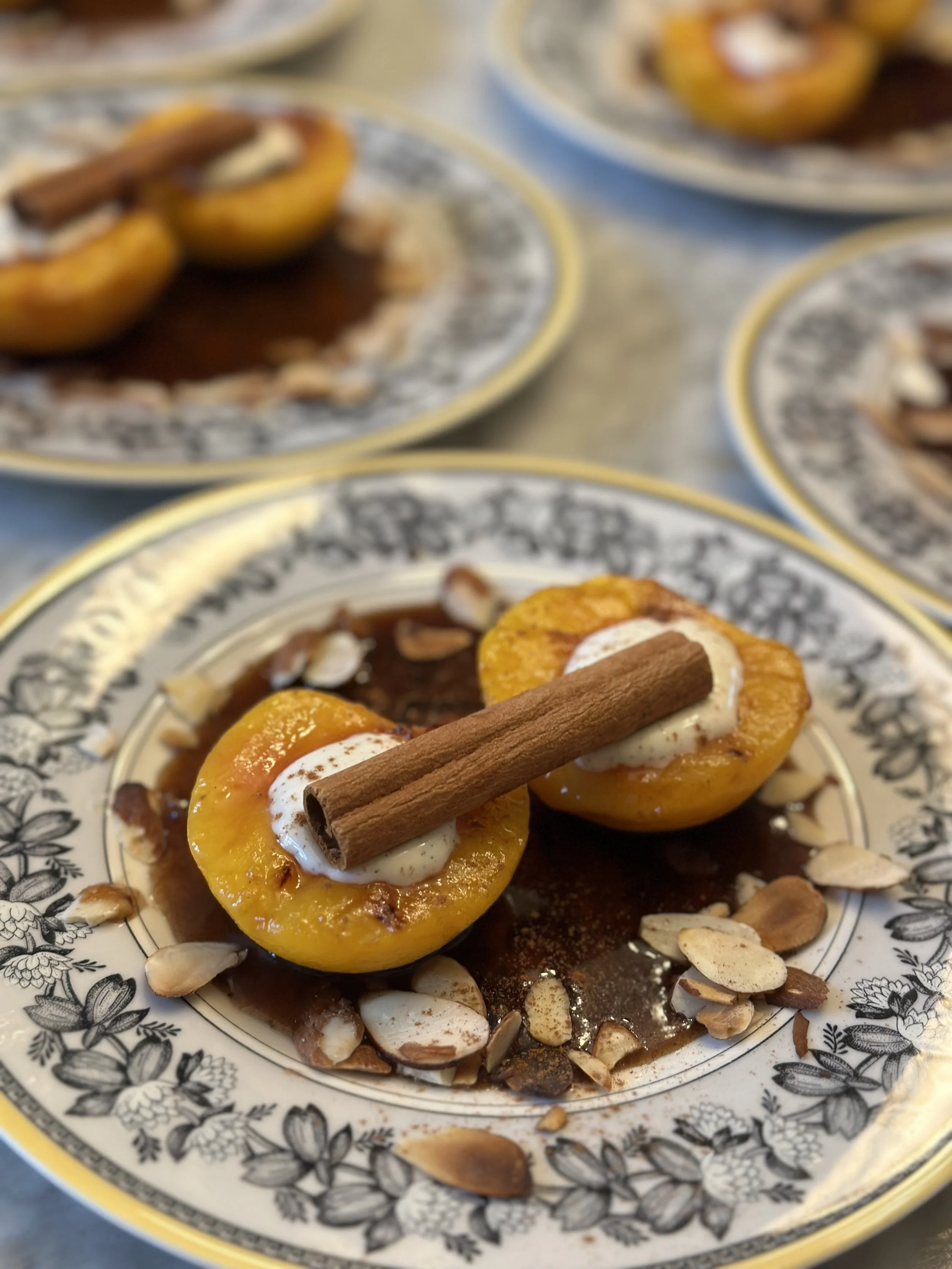 Baked peaches on a decorative plate, topped with cinnamon sticks, whipped cream, and surrounded by sliced almonds.