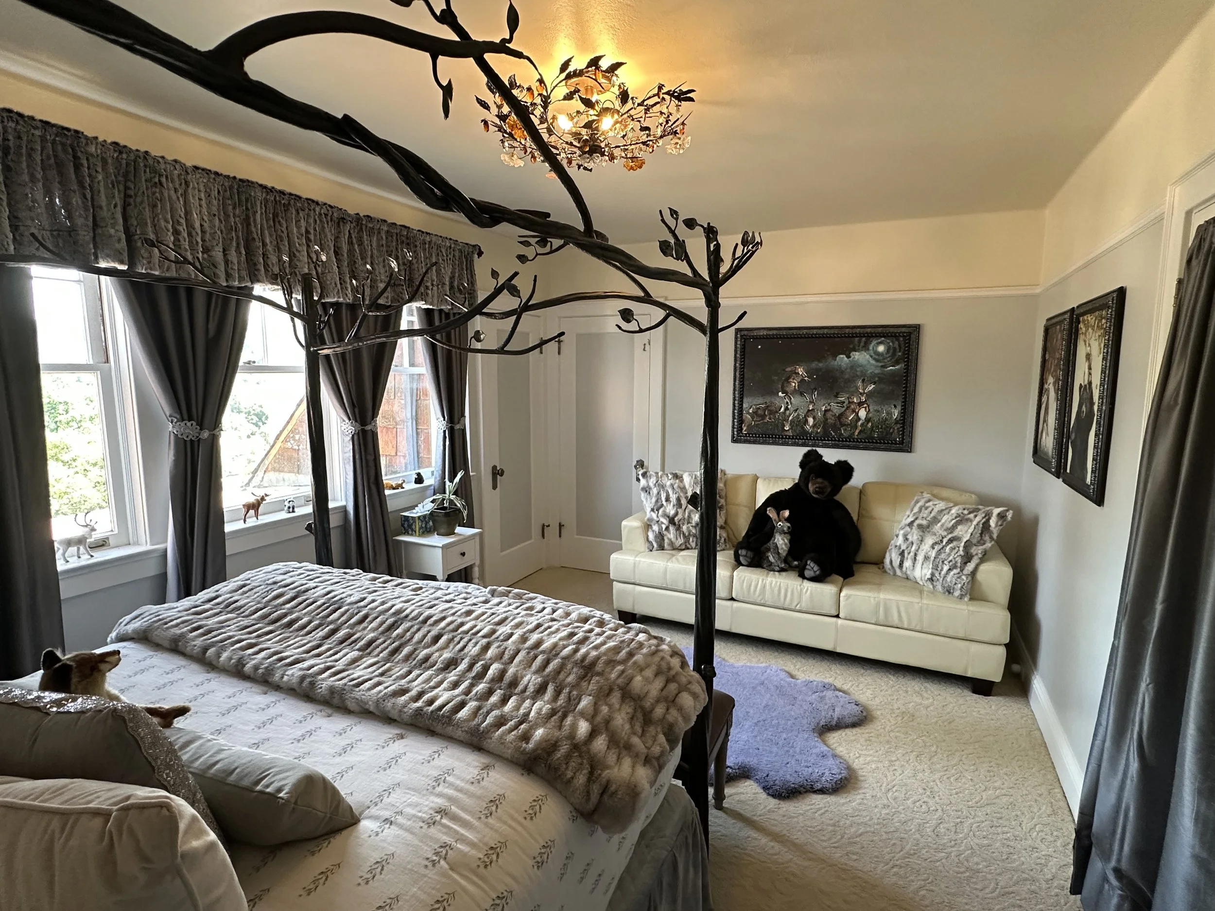 Elegant bedroom with tree branch-themed bed frame, plush bedding, large windows with dark curtains, and a sofa adorned with a teddy bear and decorative pillows. Wall art includes whimsical paintings. The room has a cozy ambiance with a fluffy area rug and soft lighting from a ceiling fixture.