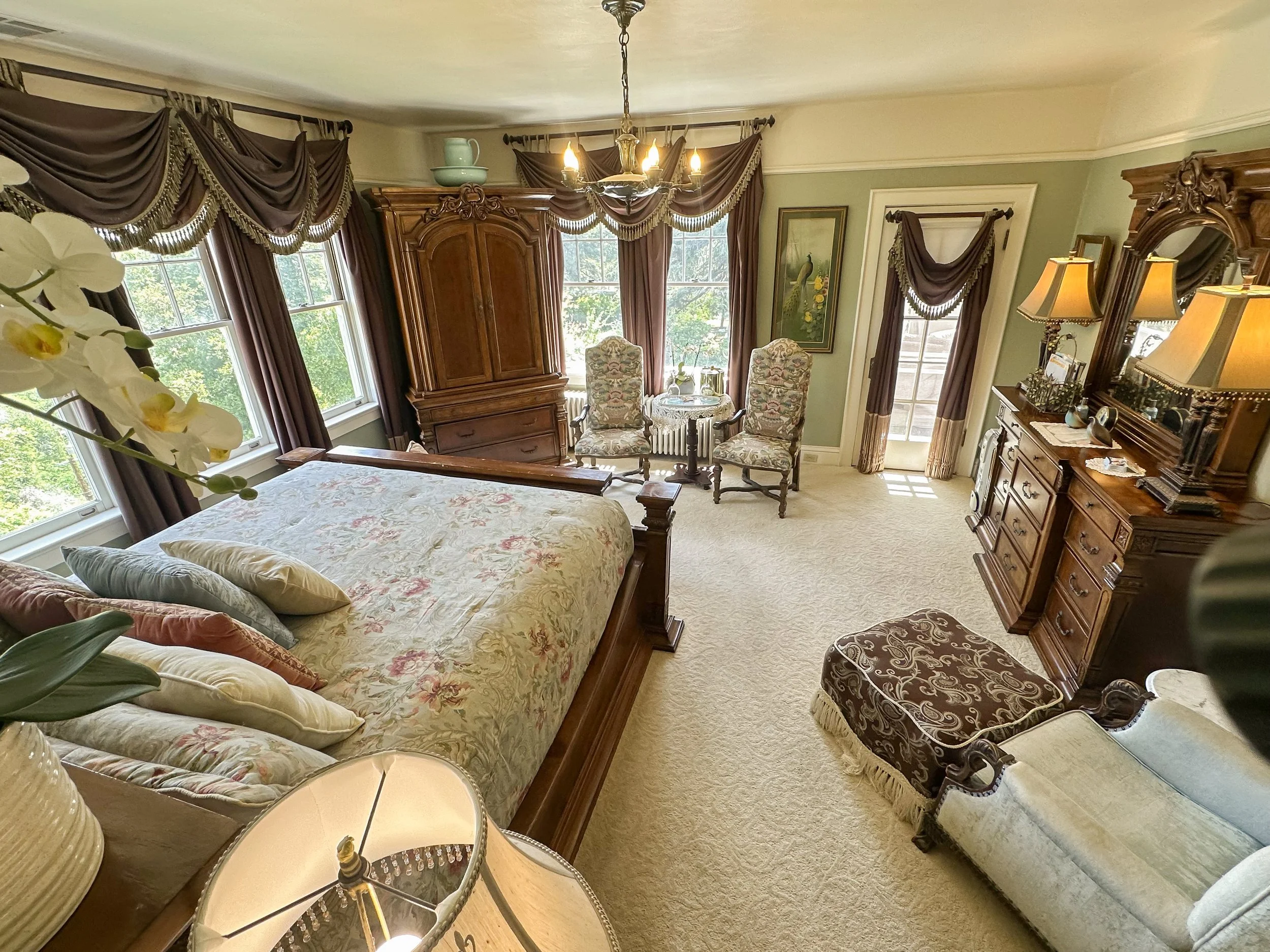 Elegant bedroom with ornate wood furniture, including a large bed with floral bedding, a dresser with matching mirror, and an armoire. The room features large windows with elaborately draped curtains, two upholstered chairs with a small round table, and stylish lamps.