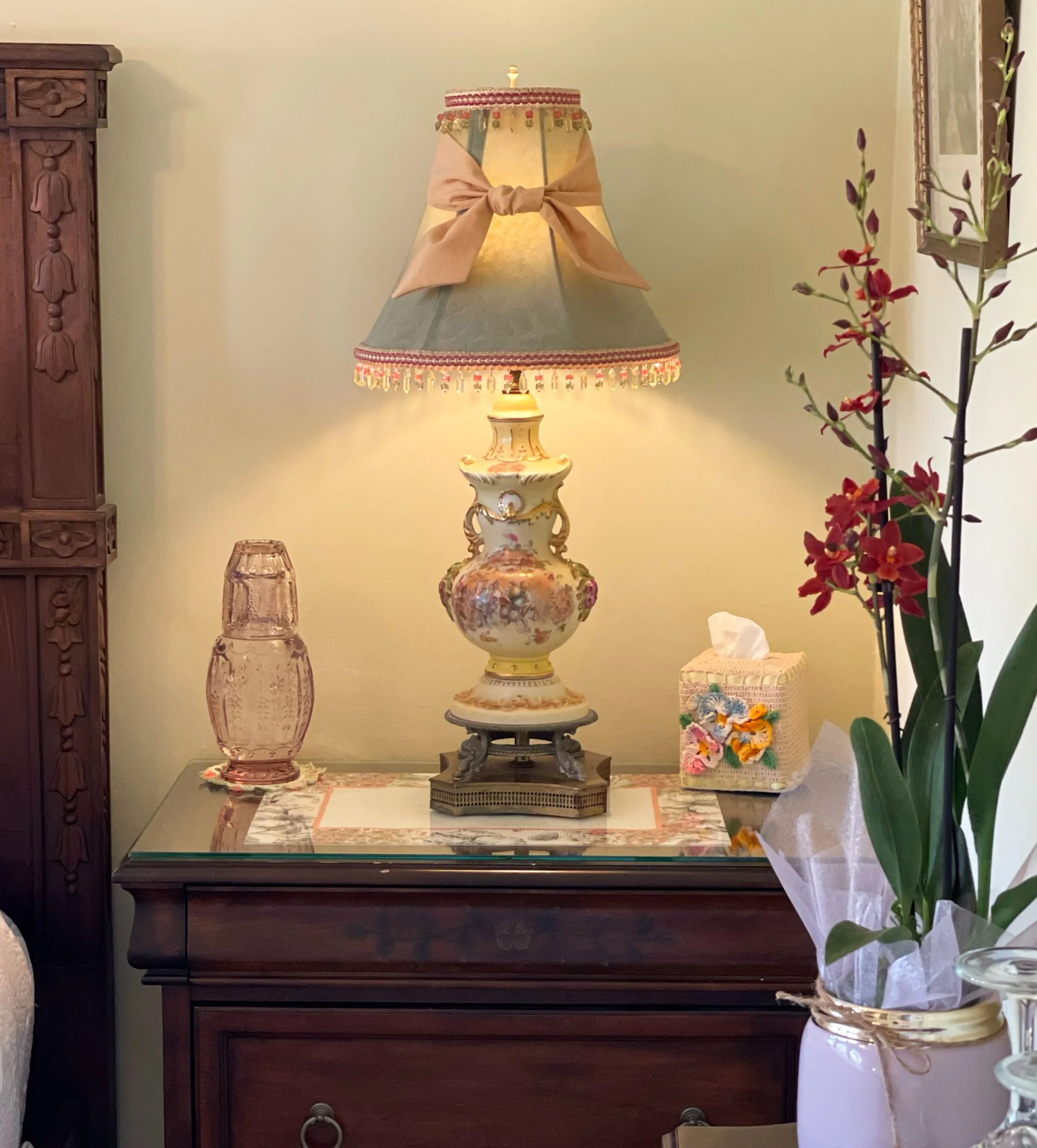 A decorative table lamp with a floral design, topped with a pink ribbon, situated on a wooden sideboard. To the left of the lamp, there is a pink glass vase, and to the right, a ornate tissue box. A large potted orchid with red flowers is on the far right, and a framed picture hangs on the wall in the background.