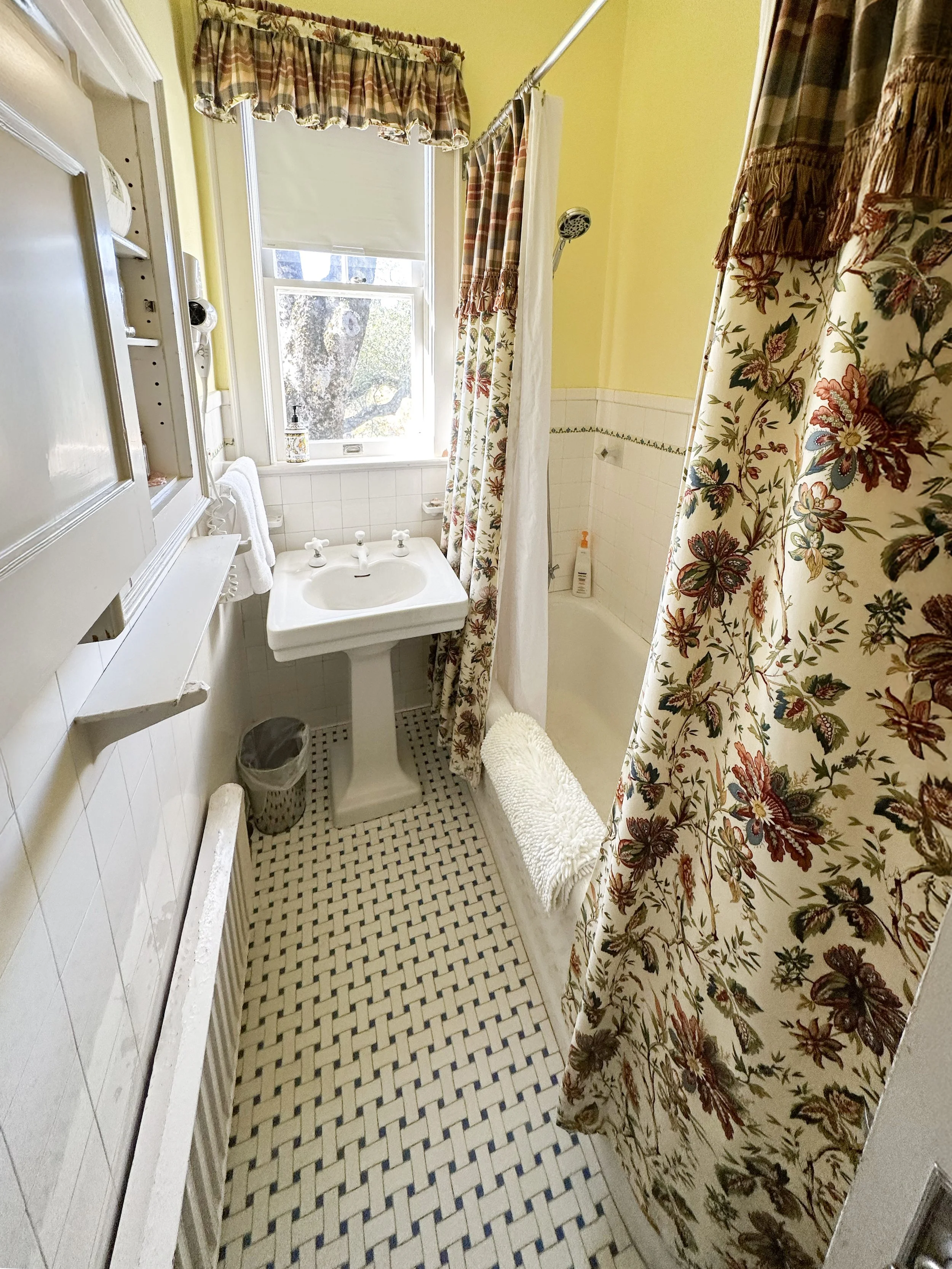 A small vintage bathroom with yellow walls, a pedestal sink, floral curtains, a window with a blind, a bathtub with a floral shower curtain, and checkered floor tiles.