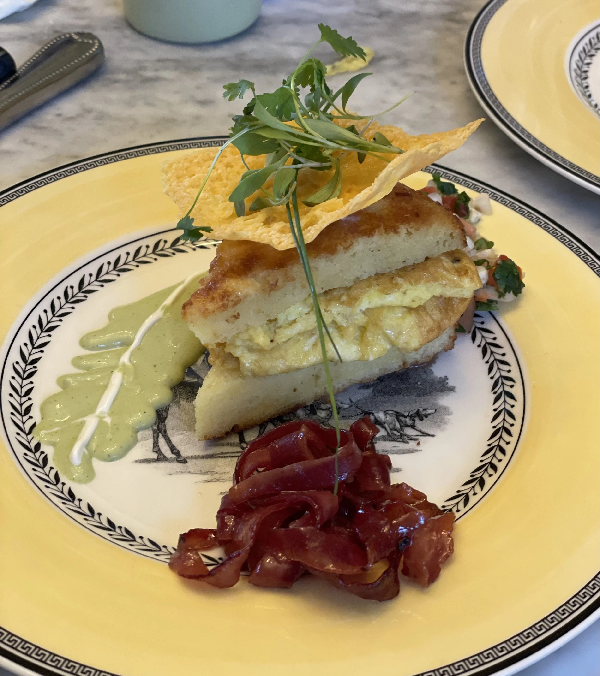 Gourmet dish featuring a sandwich with eggs, green sauce, red onion garnish, and a crispy chip, topped with herbs, on a decorative yellow plate.