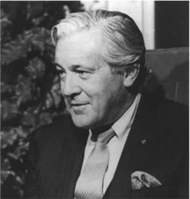 Black and white photo of a man with light-colored hair, wearing a suit, sitting indoors.
