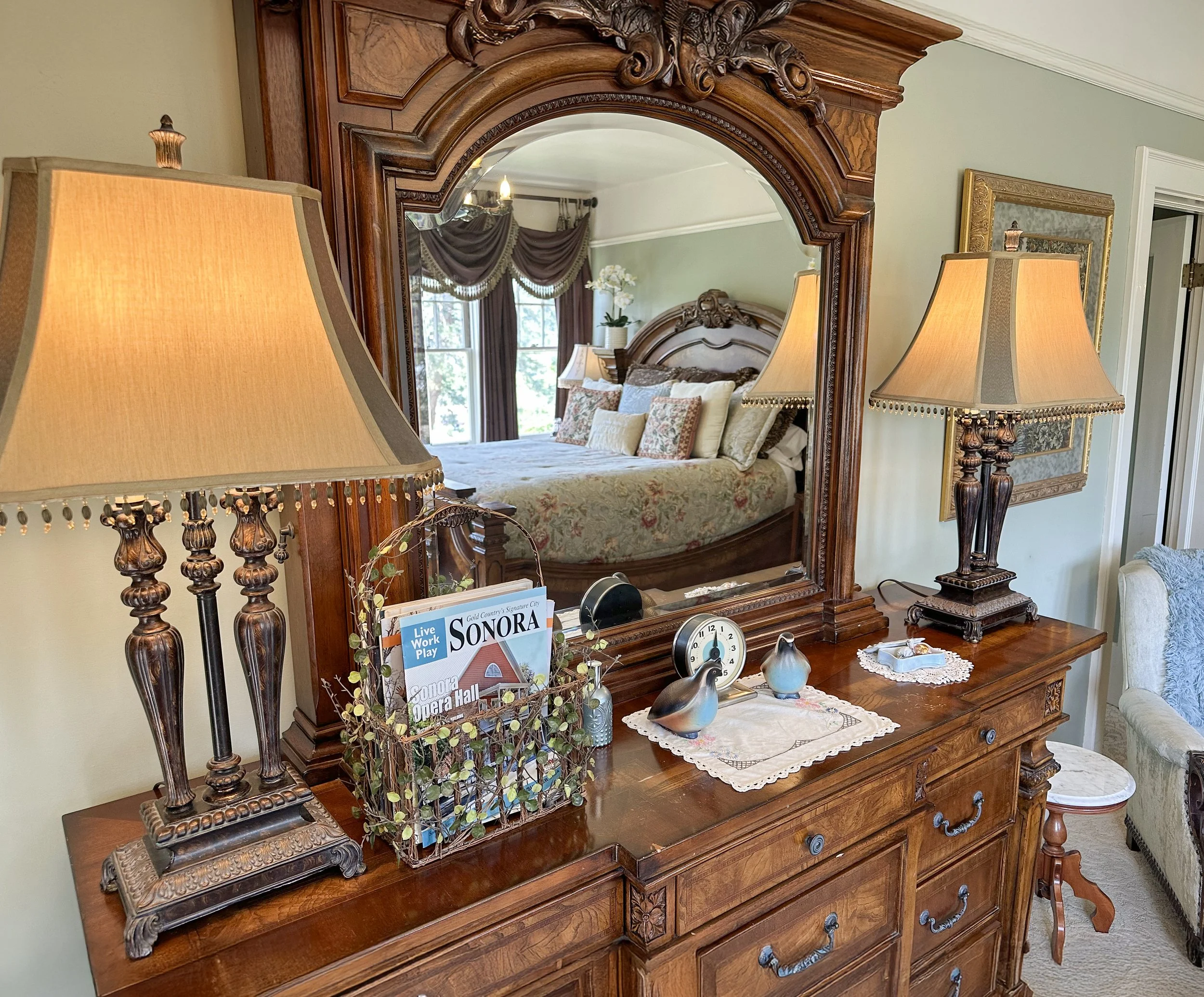 Bedroom with a large ornate wooden dresser and mirror, flanked by two decorative lamps. The dresser holds a small clock, decorative items, and a basket with magazines. The reflection shows a bed with floral bedding, pillows, and a window with elegant curtains.