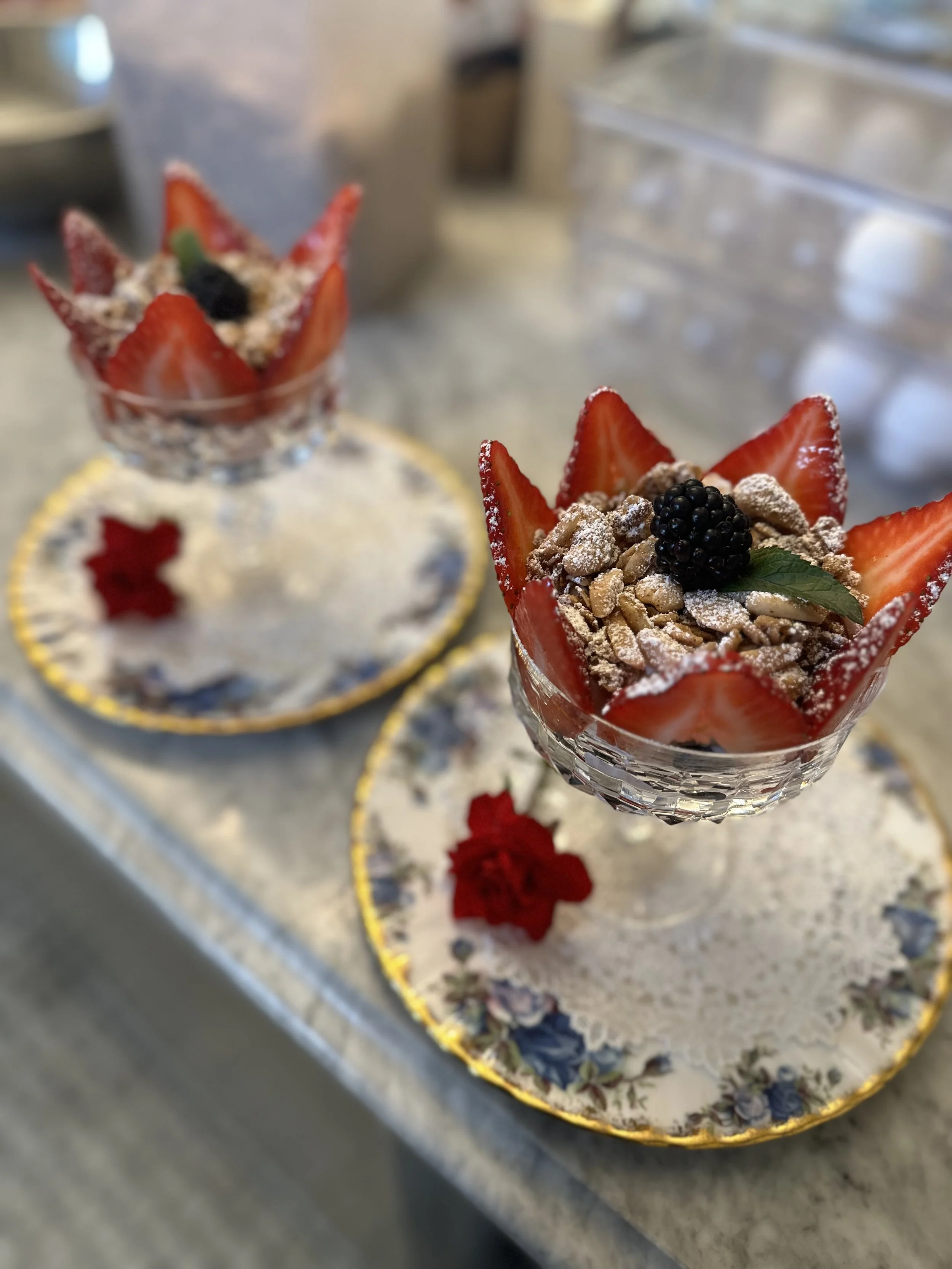 Two elegant desserts in crystal glasses, garnished with sliced strawberries, blackberries, and powdered sugar, served on floral-patterned plates with red flower decorations.