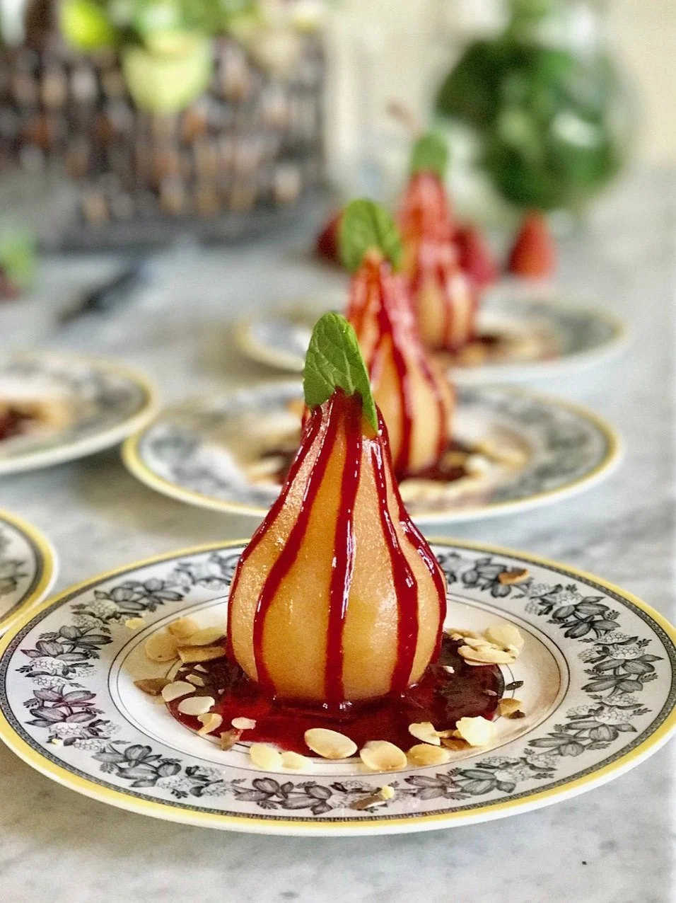 Poached pear dessert with red sauce drizzle on decorative plate, garnished with mint leaf and sliced almonds.