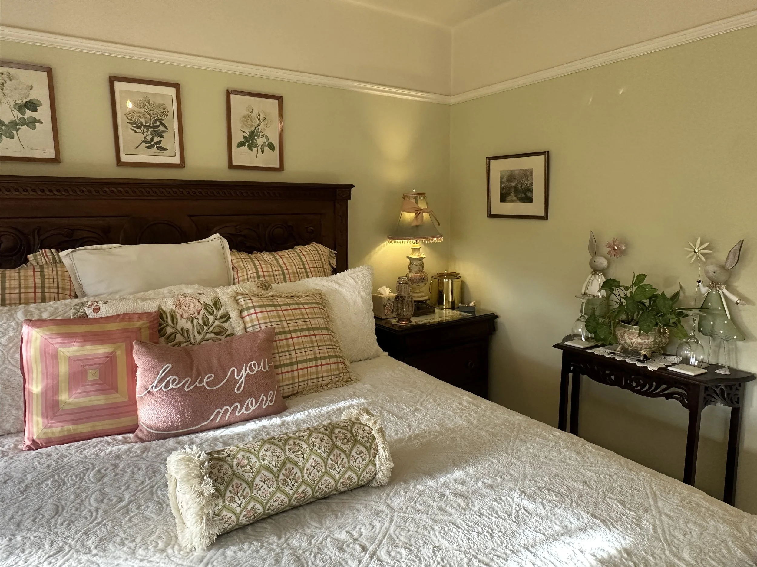 Cozy bedroom with decorative pillows on a bed, floral prints on the wall, a lamp on a nightstand, and a small table with plant decorations and bunny figurines.