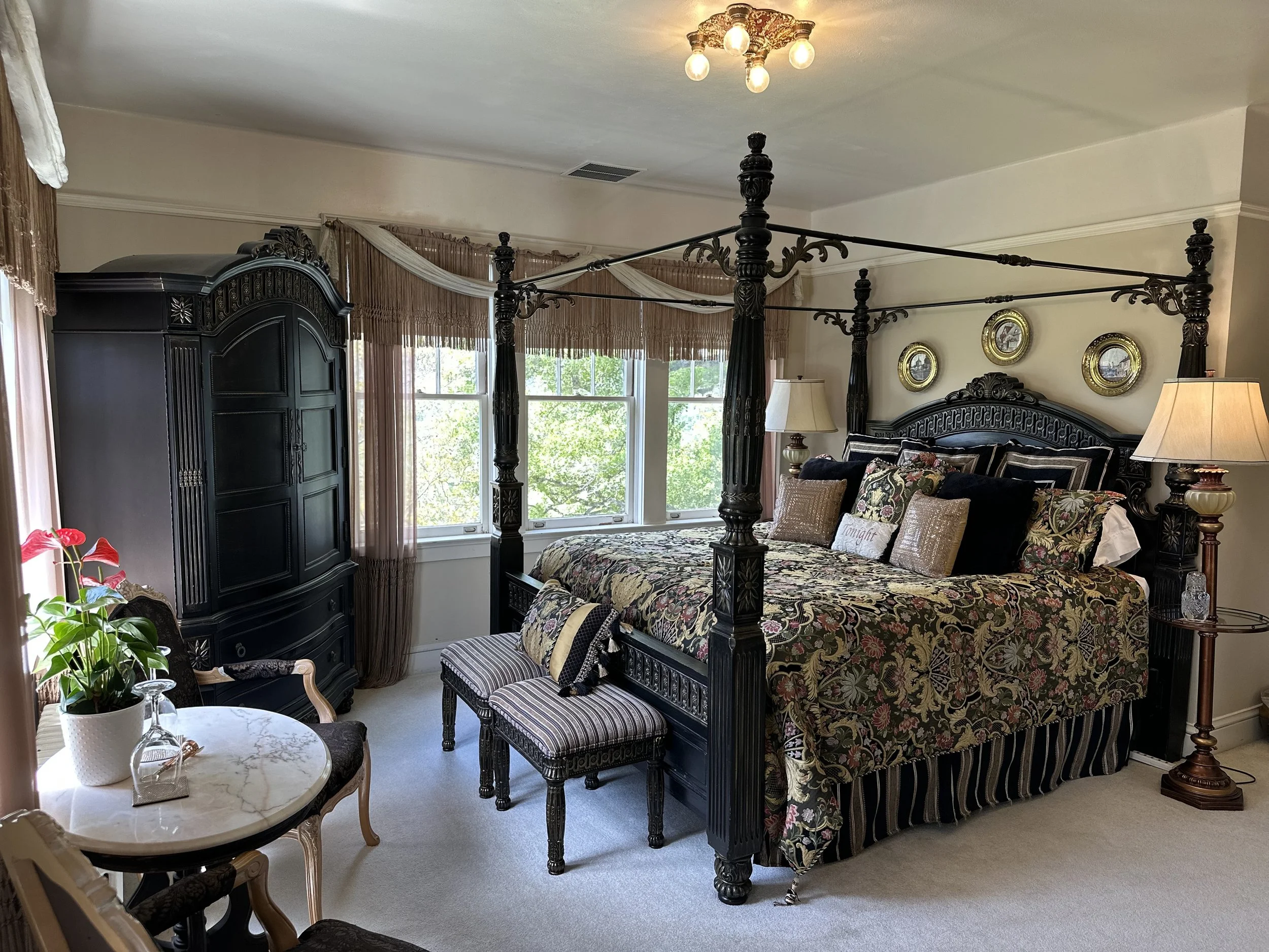 Luxurious bedroom featuring ornate black furniture, including a canopy bed with patterned bedding and throw pillows, a matching bench, and an armoire. Two side lamps provide ambient lighting. The room has a small table with chairs, decorated with a plant and glass items, positioned near windows with sheer curtains.