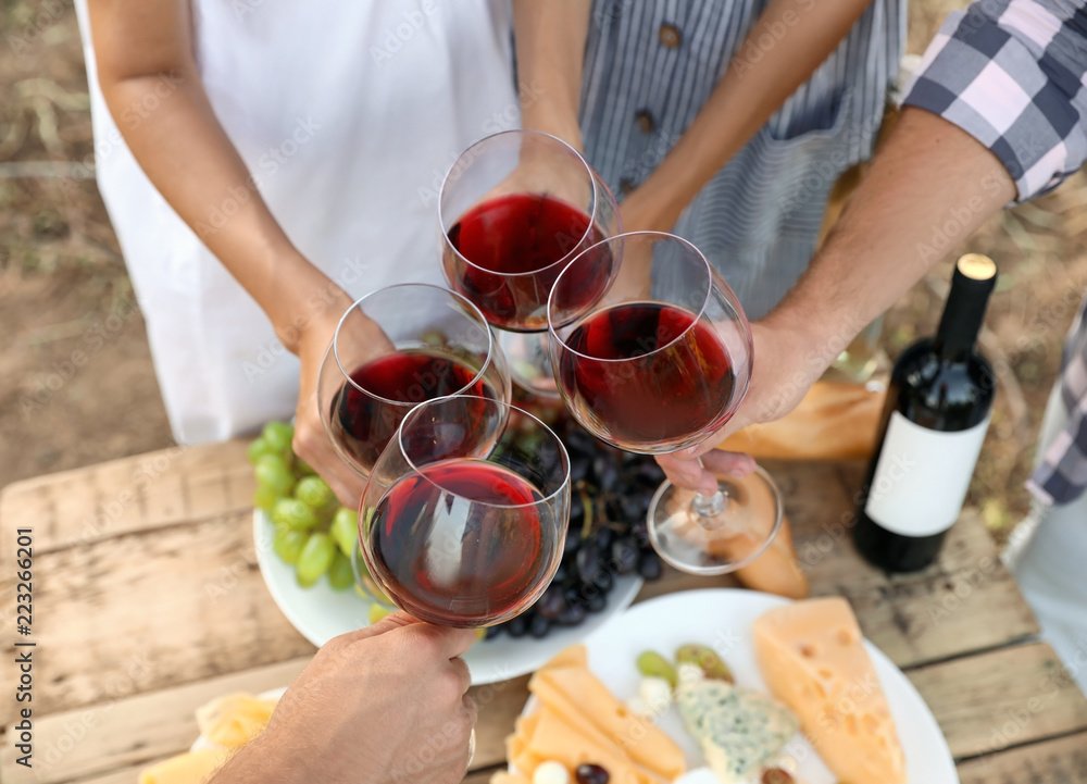 Four people clinking glasses of red wine above a rustic wooden table with grapes, cheese, and a bottle of wine.