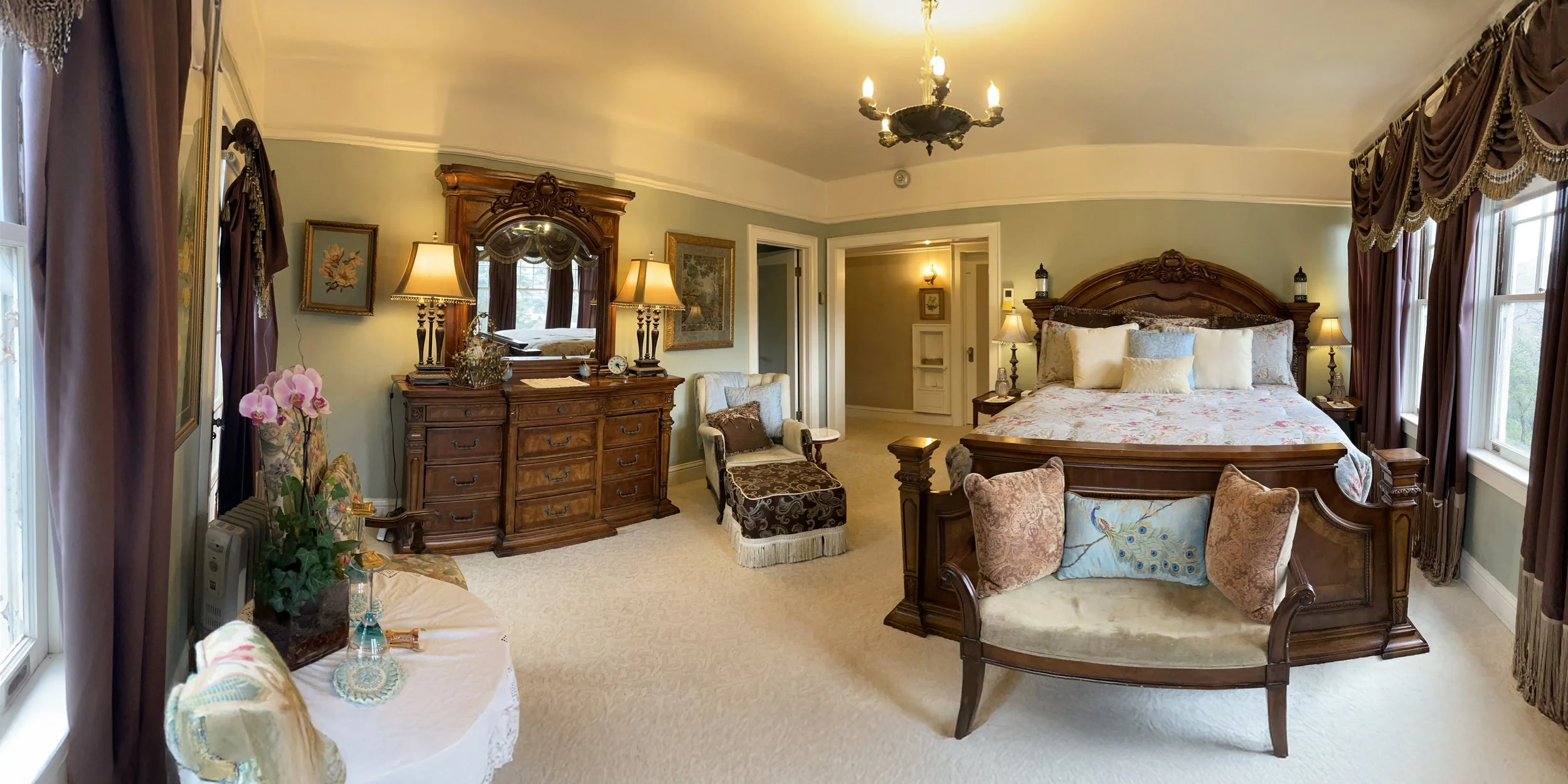 Luxurious bedroom with elegant wooden furniture, large ornate bed, decorative pillows, and floral quilt. Room features chandeliers, large windows with heavy drapes, a dresser and mirror, armchair with ottoman, and a small table with orchids.